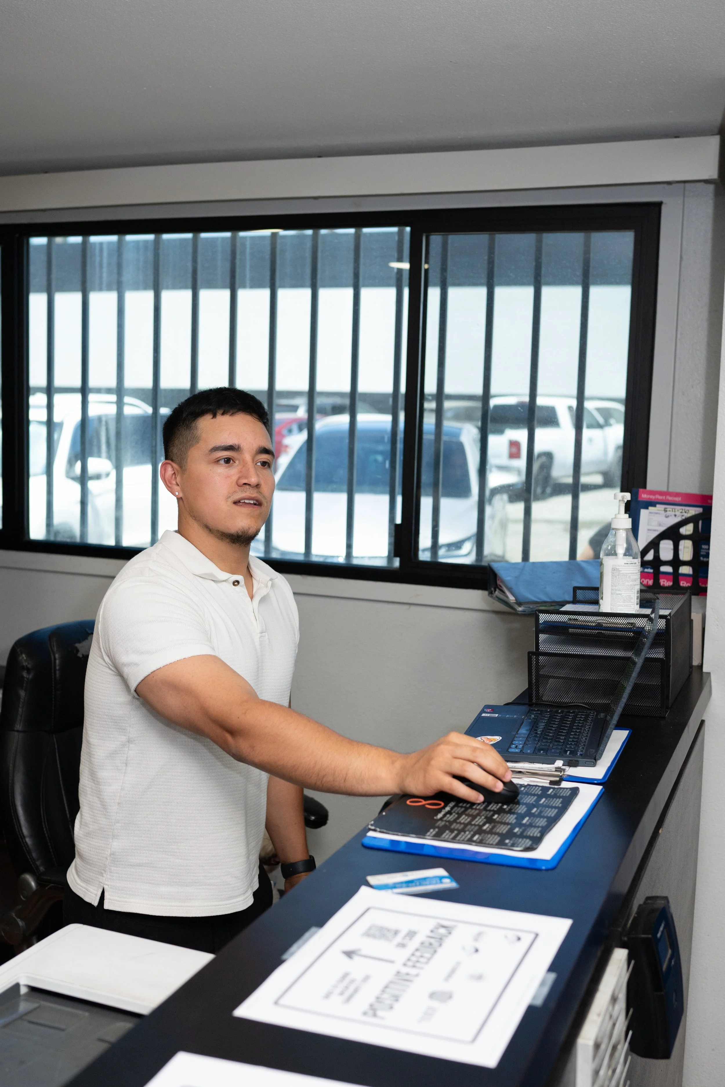 A young man in a white polo shirt sitting at a desk in an office, with a computer mouse in his right hand and a laptop in front of him. There are various office supplies and documents on the desk. A large window with black bars and vehicles outside i