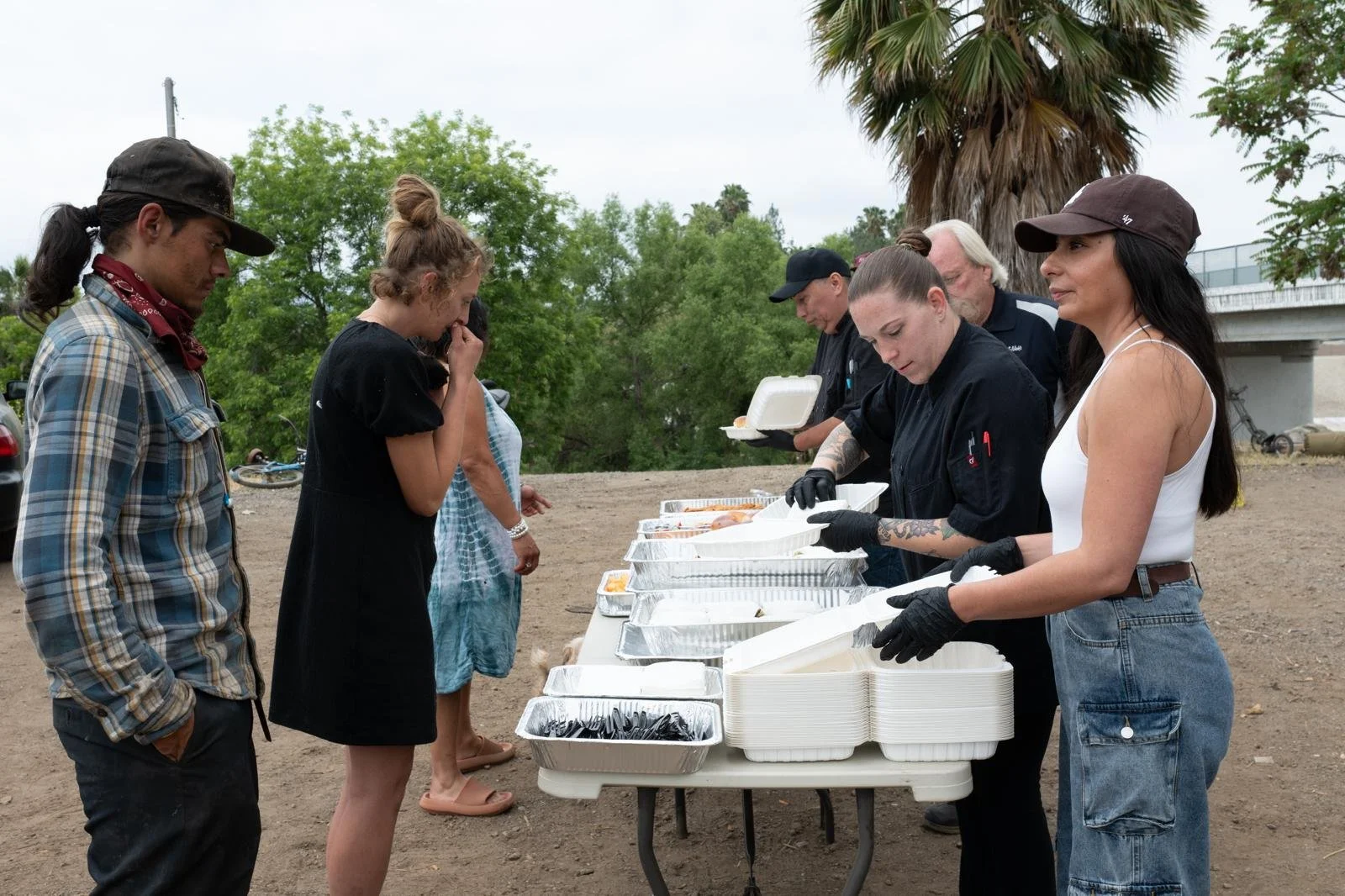 People receiving food from a catering line outdoors with trees and a bridge in the background.