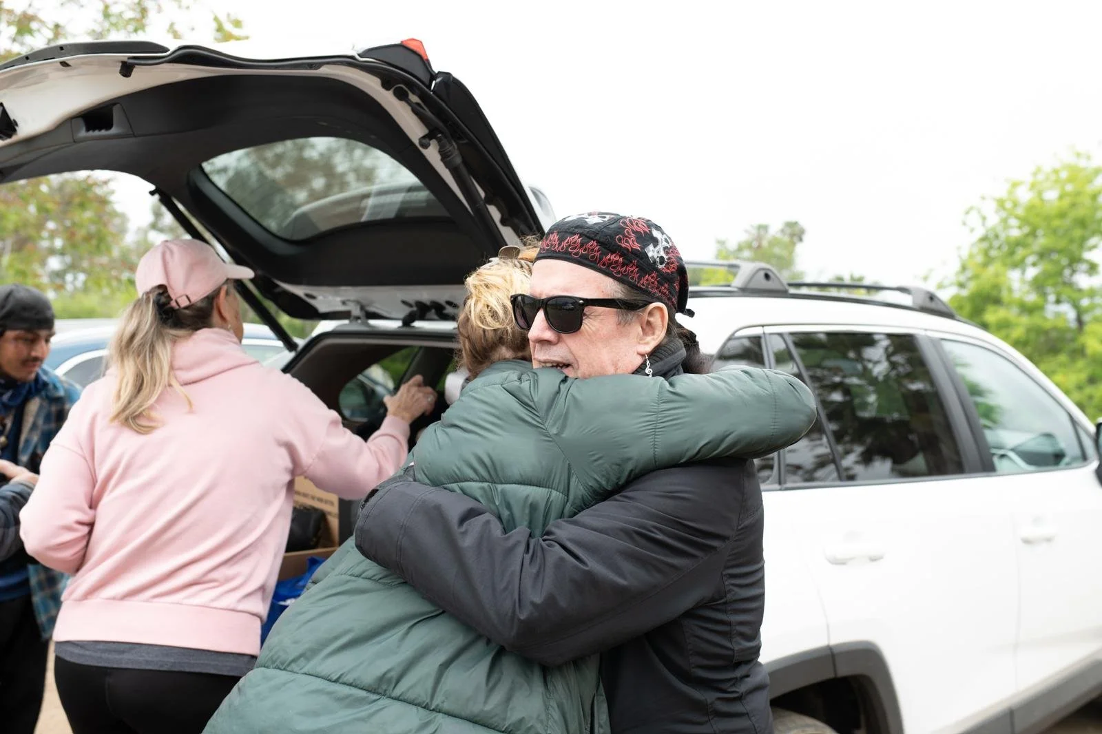 People hugging in front of an open car trunk, with others nearby, outdoors with trees in the background.