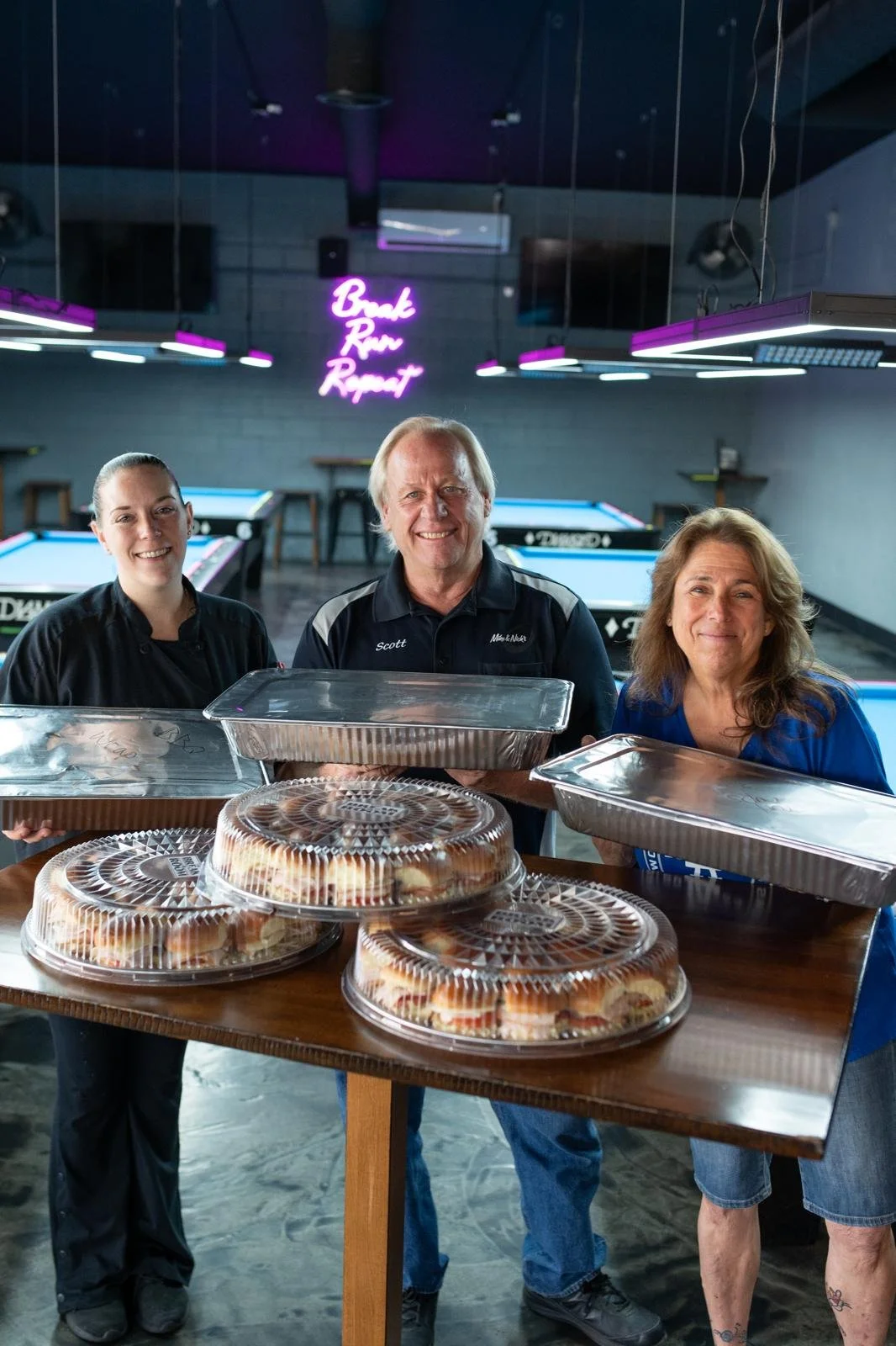 Three people standing behind a table with trays of baked goods in a pool hall with pool tables and a neon sign that says 'Break Run Repeat' in the background.