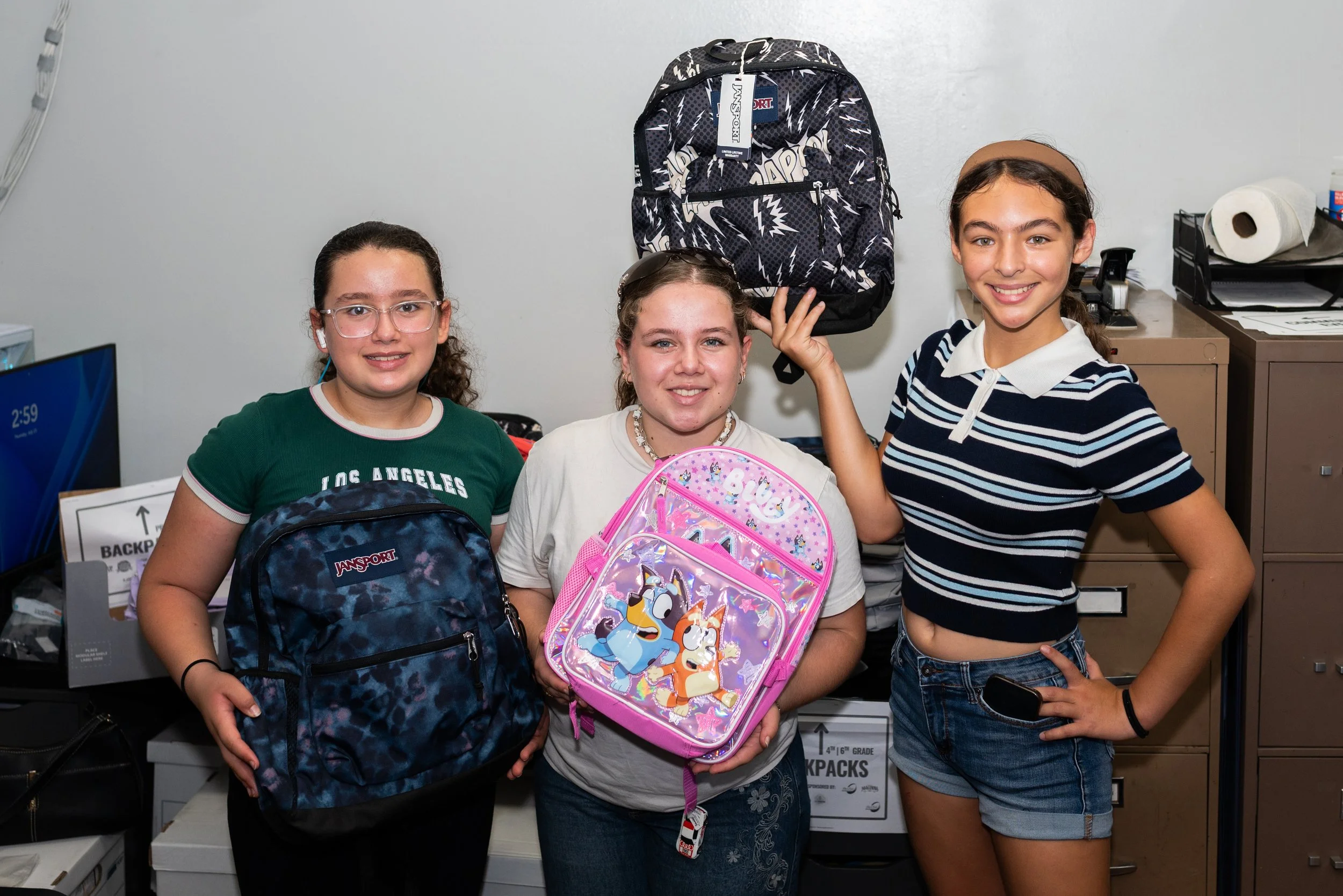 Three young girls are standing together in an office environment, each holding or wearing backpacks, smiling at the camera.
