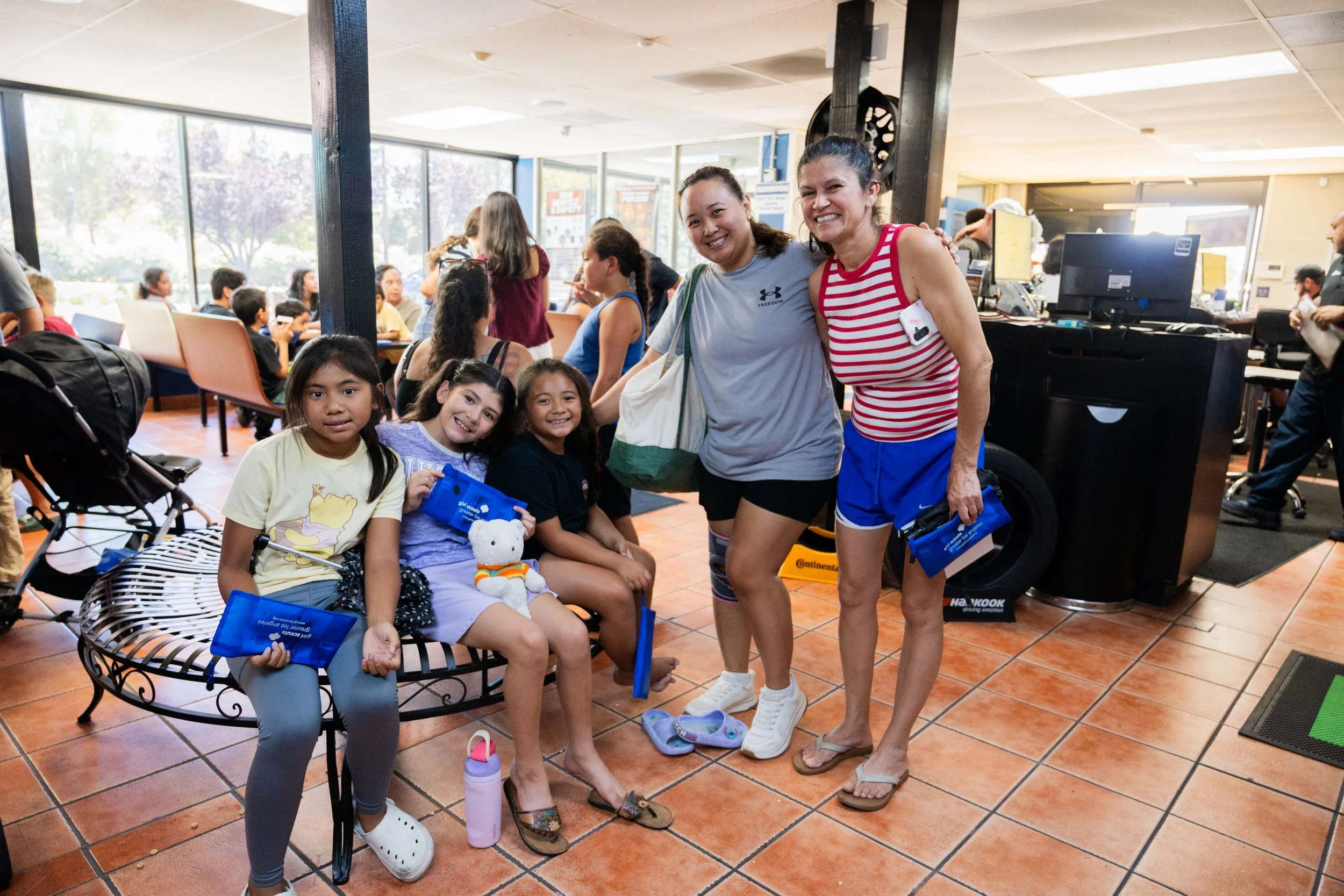 A group of children and two women smiling inside a room, with children seated in the background. The room has large windows and various equipment. The children are holding blue bags and some have toys, while the women are standing and smiling, one we