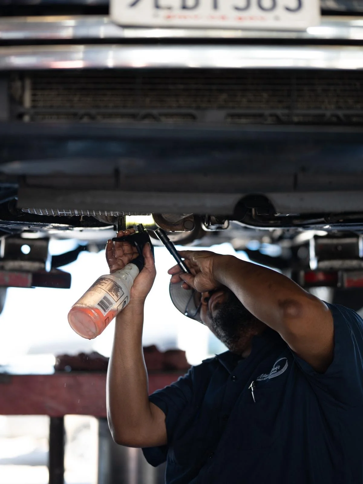A mechanic working underneath a vehicle, holding a spray bottle and a tool, inspecting or repairing a part of the car's underside.
