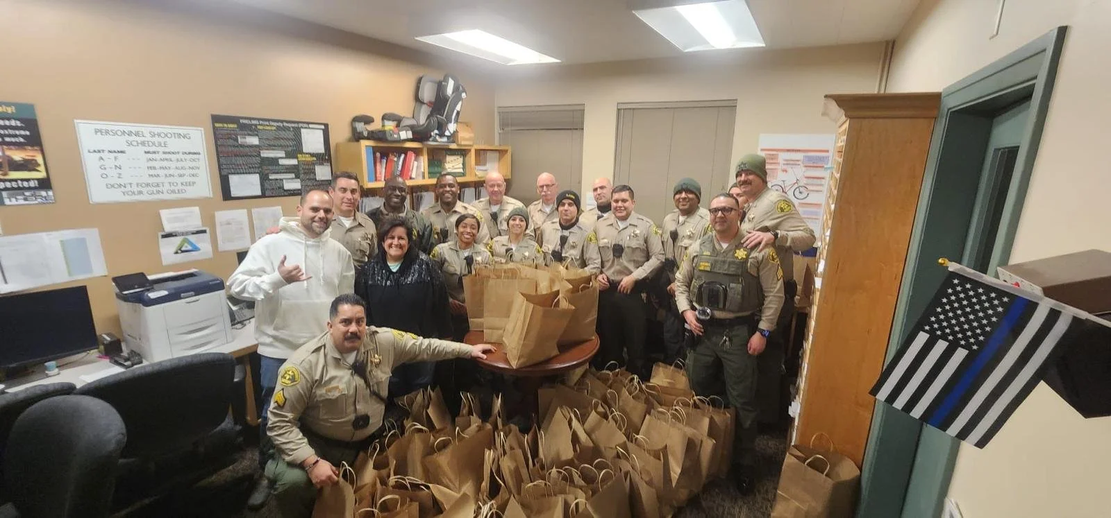 Group of police officers and civilians in an office room with gift bags on a table and on the floor, a police flag on the wall, and various posters and shelves in the background.