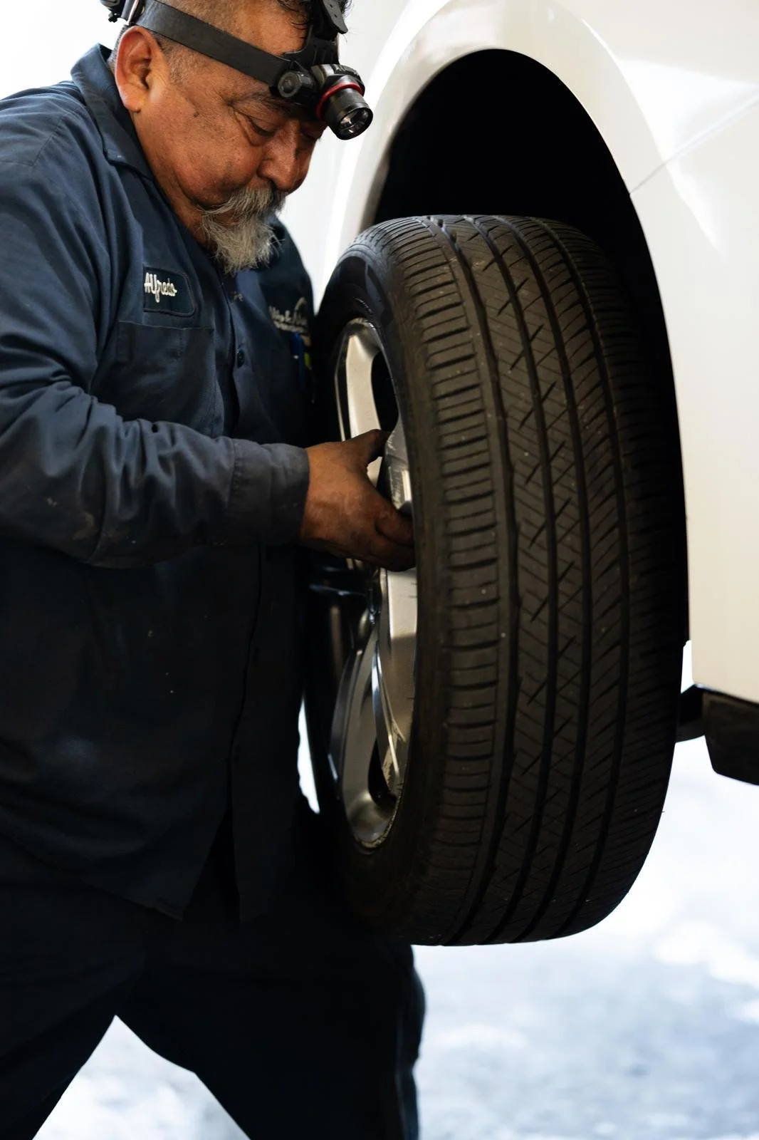 A mechanic changing a tire on a white vehicle, wearing a black uniform with a name tag, and a headlamp for illumination.