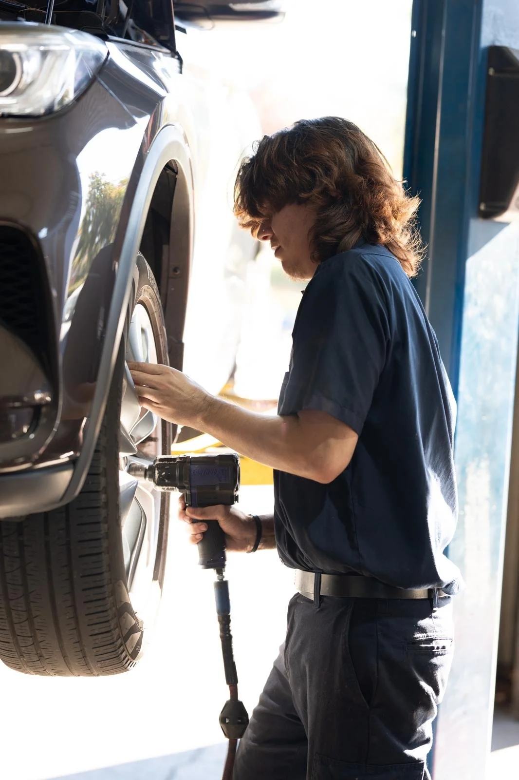 A mechanic working on a car lifted in a garage, using a power drill to work on the wheel.