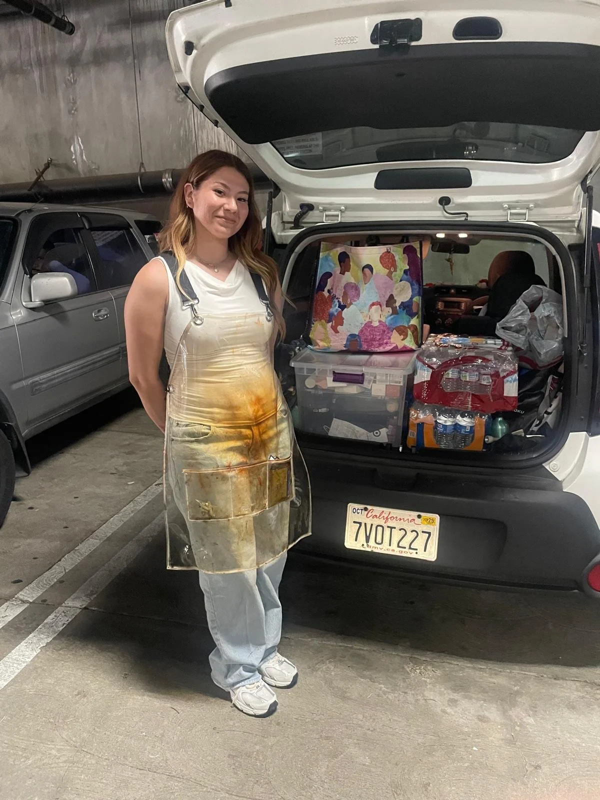 A woman standing in a parking garage next to an open trunk of a white car filled with various bags and supplies. She is wearing a clear apron with paint stains and casual clothes. The car has a California license plate reading '7VOT227'.