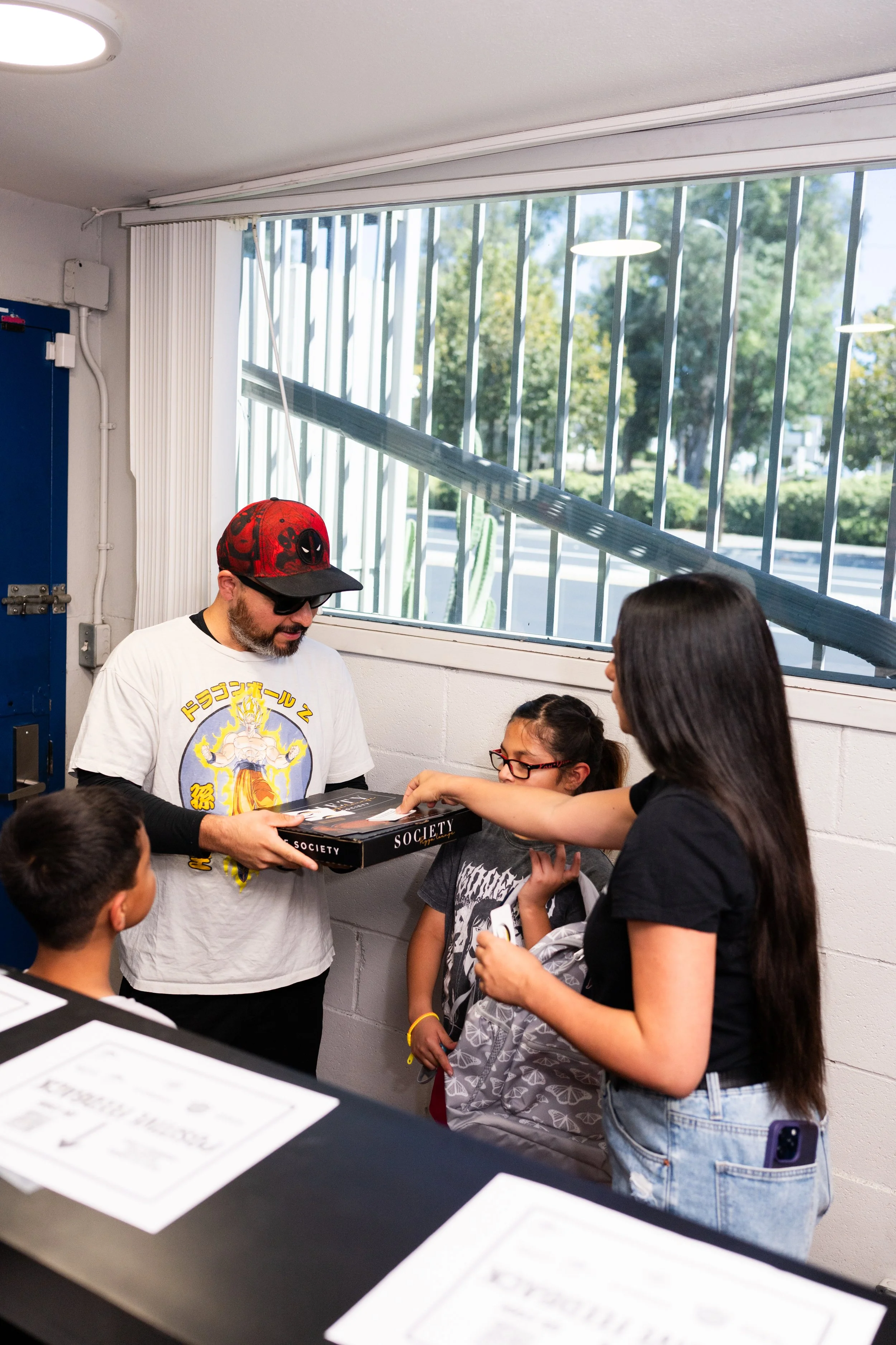 A man wearing sunglasses and a Deadpool hat hands a board game titled 'Society' to a girl wearing glasses and a grey shirt with butterfly designs, while other children and a woman observe.