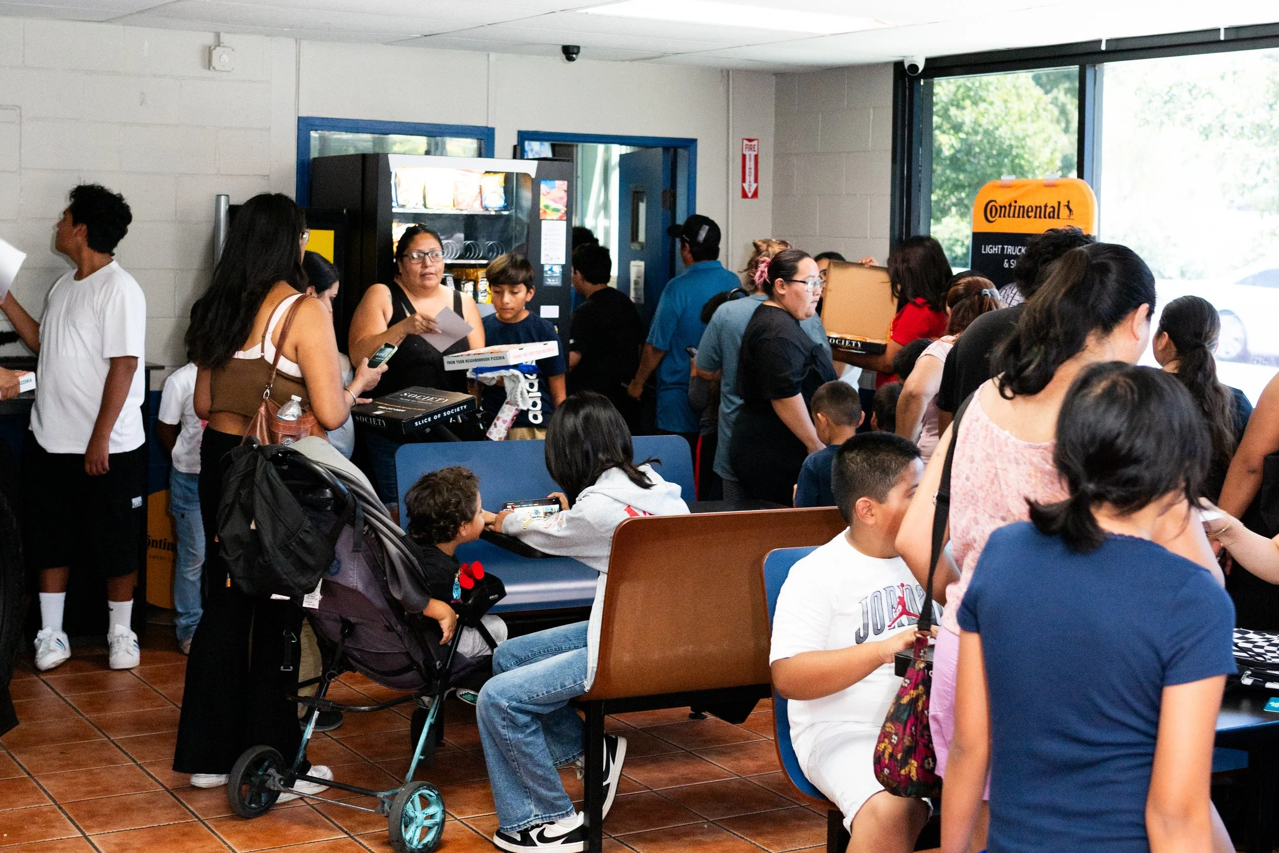 Many people, including children and adults, are standing and sitting in a busy waiting area inside a building. There is a vending machine near the wall, and some people are using their phones or talking. A stroller is also visible. The scene appears 