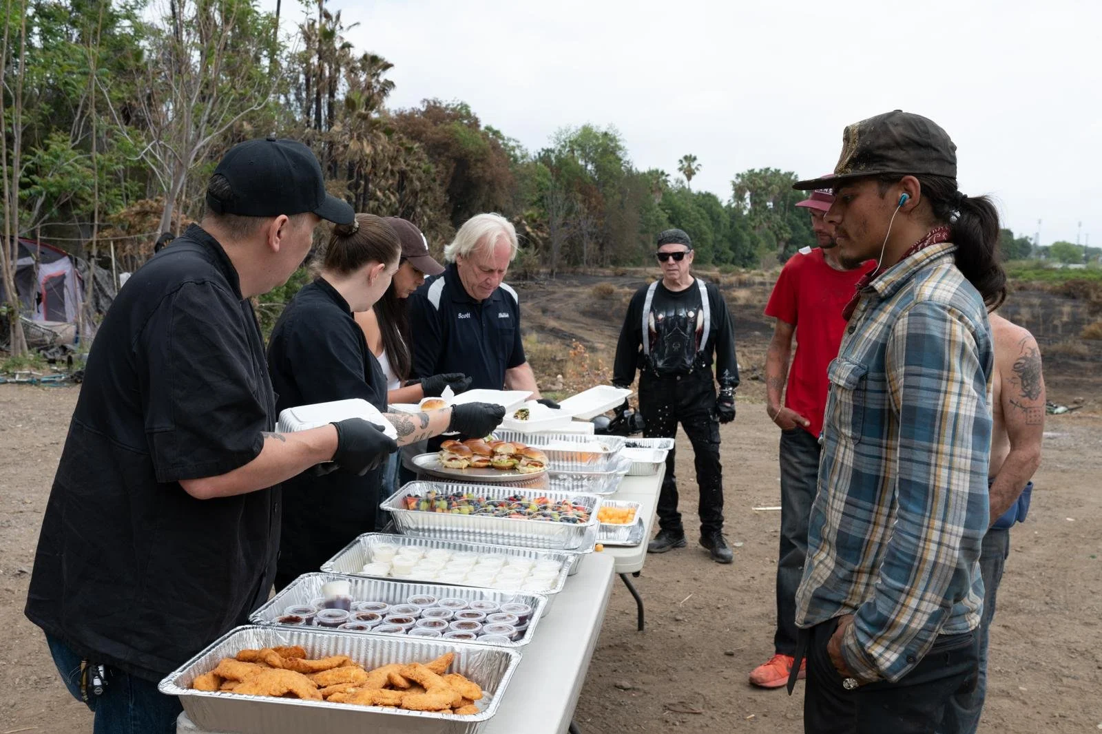 People standing in line at an outdoor food table, with some serving and others waiting, in a natural setting with trees and an overcast sky.