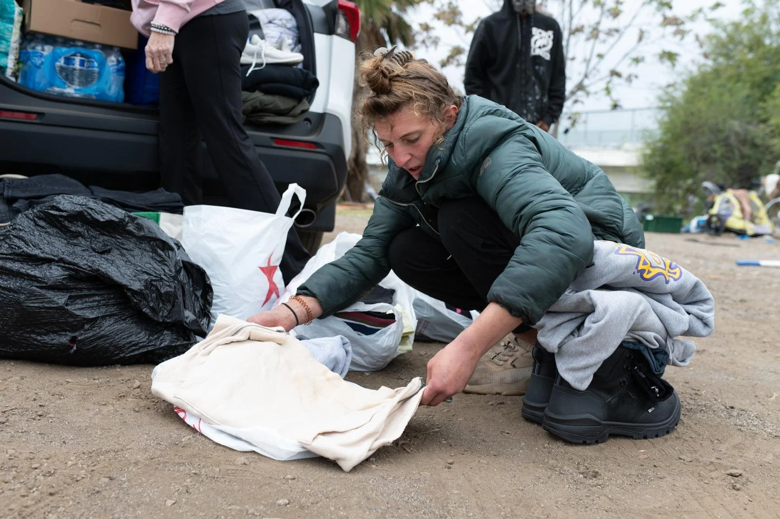 A woman kneeling on the ground, organizing clothes and bags from an open vehicle trunk during daytime.