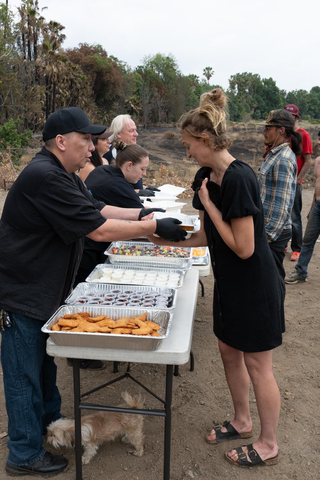 People at a food distribution event outdoors, with trays of fried foods, sauces, and desserts on a table, as a woman in a black dress receives food from a server. A small dog is under the table.