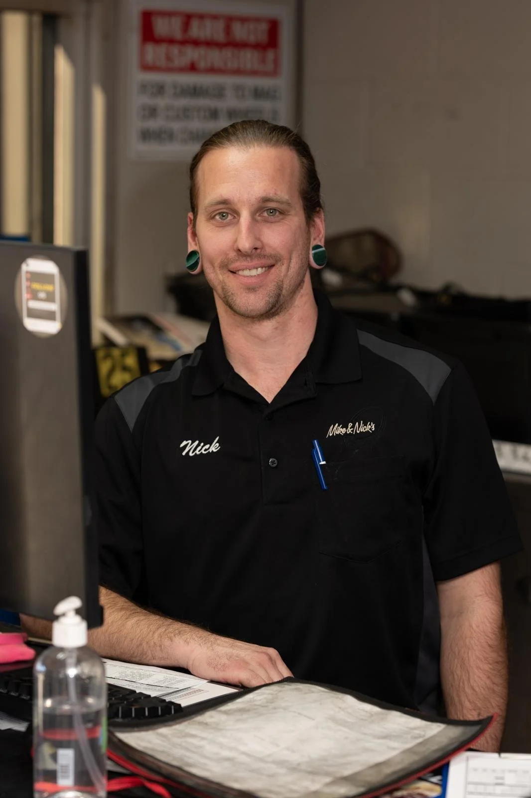 A man with long hair, teal earrings, and a beard, smiling and wearing a black shirt with a name tag that says 'Nick', sitting at a desk with a computer, on a work counter in a service or retail environment.