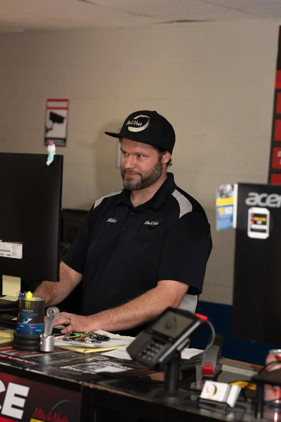 A man with a beard wearing a black cap and black shirt with gray accents, working at a counter with various items including a computer monitor, a phone, and miscellaneous objects.