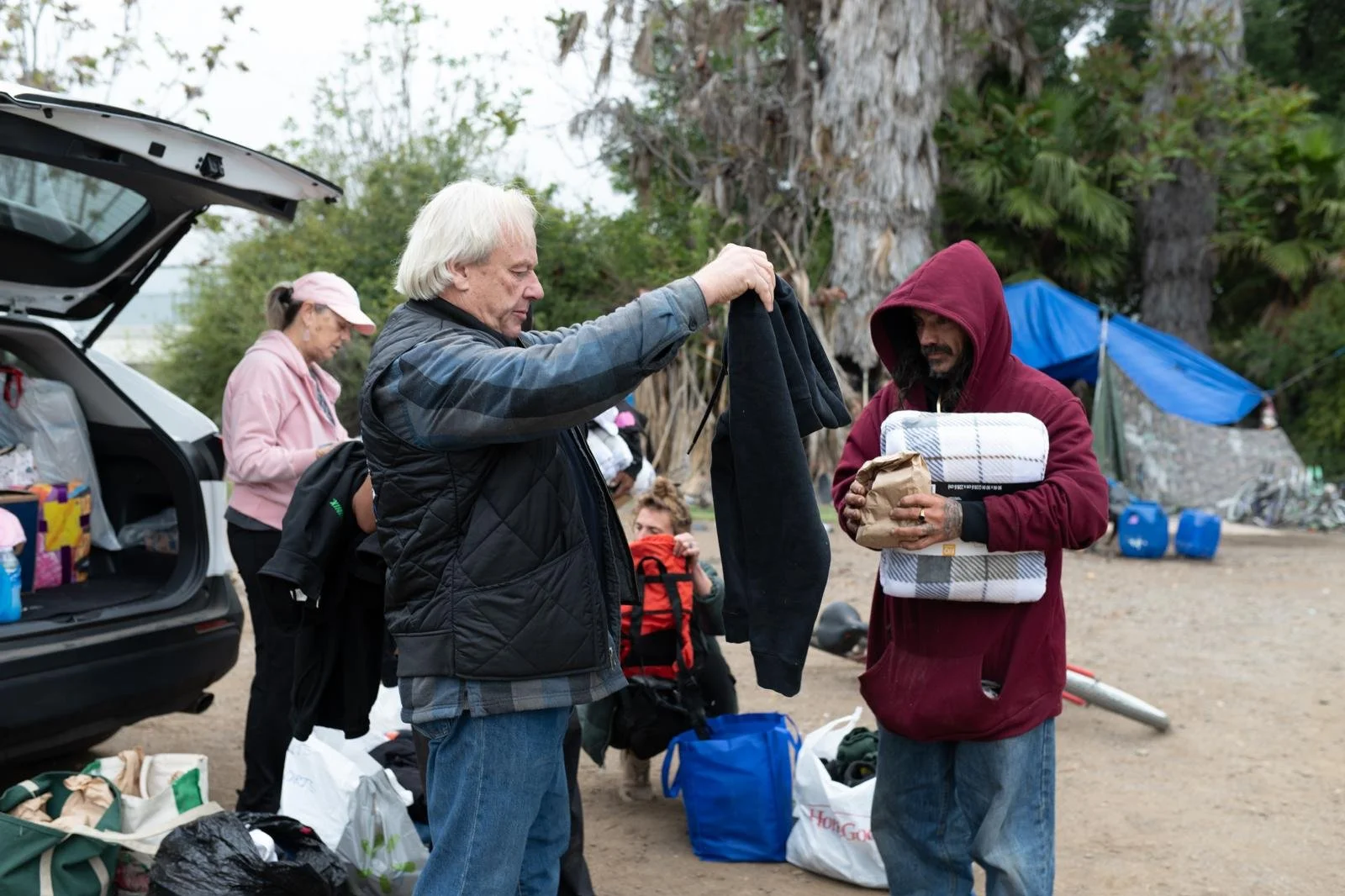 Two men exchange clothing and items at a campsite, with a group of people and camping gear in the background outdoors.
