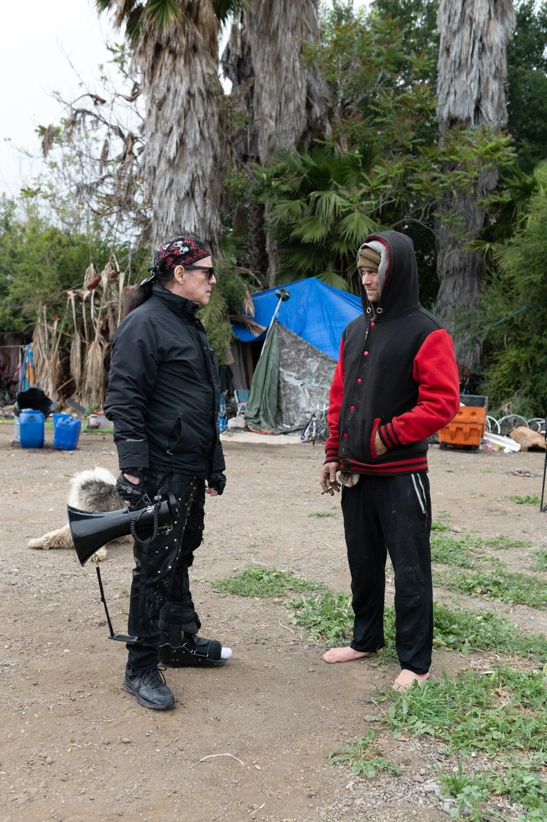 Two men standing outdoors, having a conversation. One man is dressed in black with a megaphone and a dog leash; the other man is wearing a black and red jacket with a hood, barefoot. There are large trees and makeshift tents or shelters in the backgr