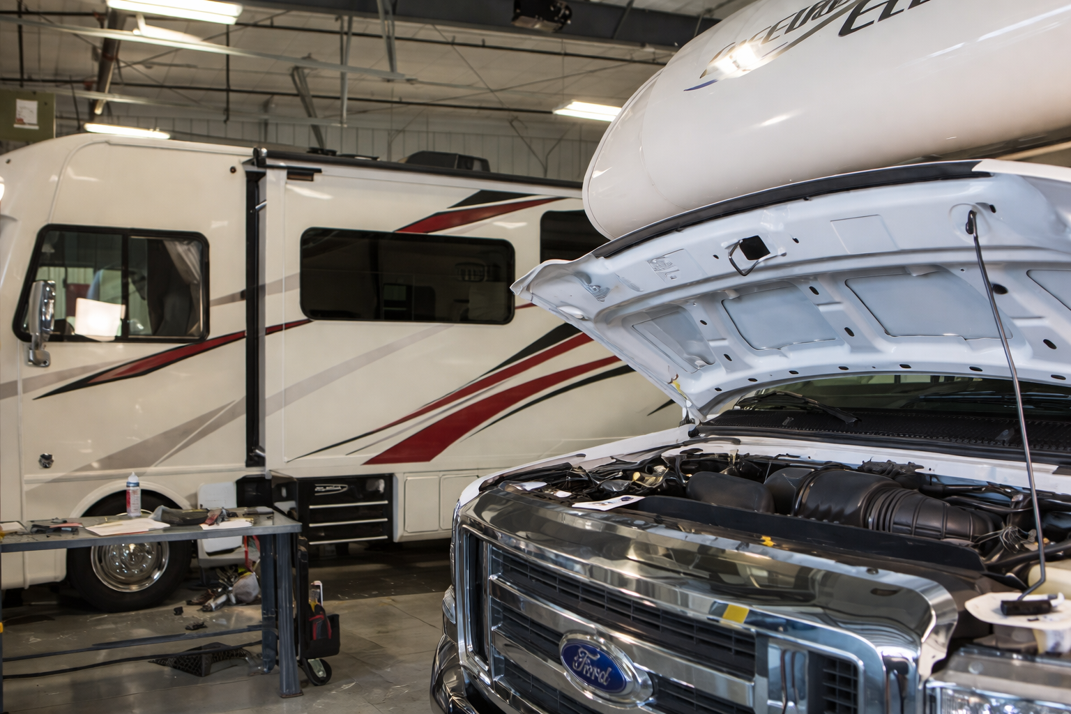 A white Ford truck with its hood open in an auto repair shop, with a recreational vehicle (RV) parked behind it.