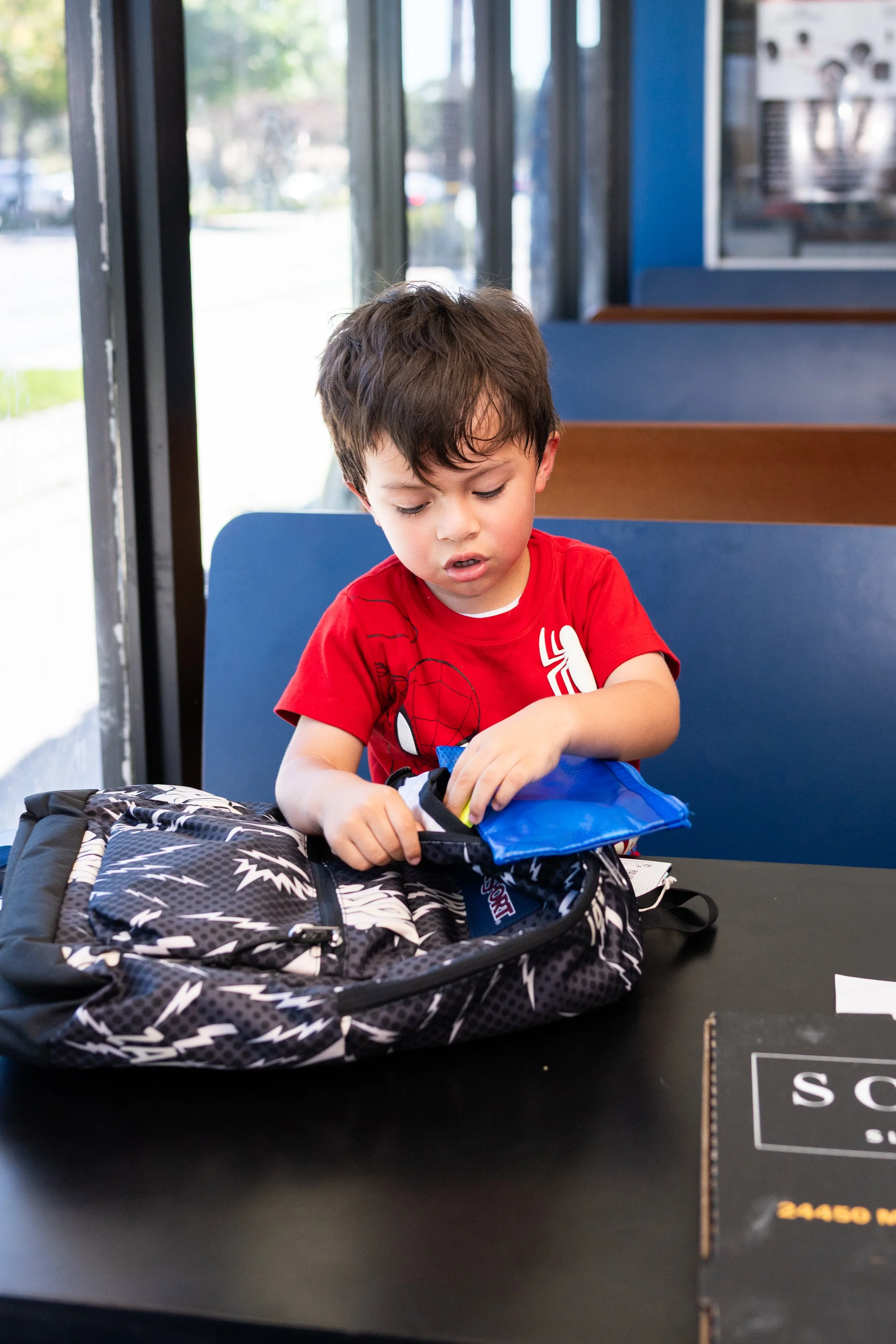 A young boy with brown hair in a red shirt sitting at a table, looking at a blue item inside a black and white backpack with lightning bolt designs, near an airport waiting area.