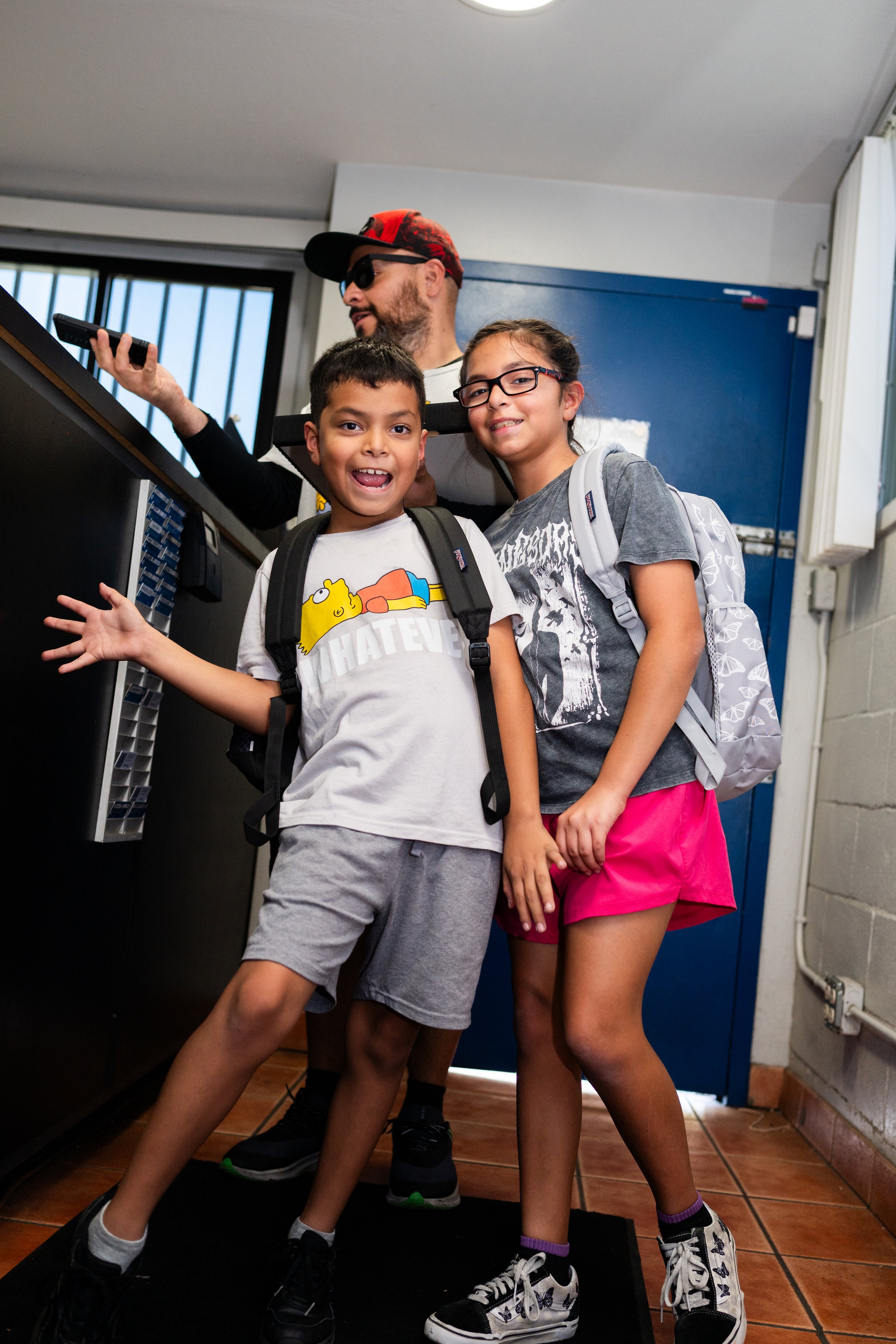 Two kids, a boy and a girl, posing playfully in a room with a man in the background. The boy is wearing a white T-shirt with a cartoon character and gray shorts, smiling and pointing, while the girl is wearing a gray T-shirt with a graphic, pink shor