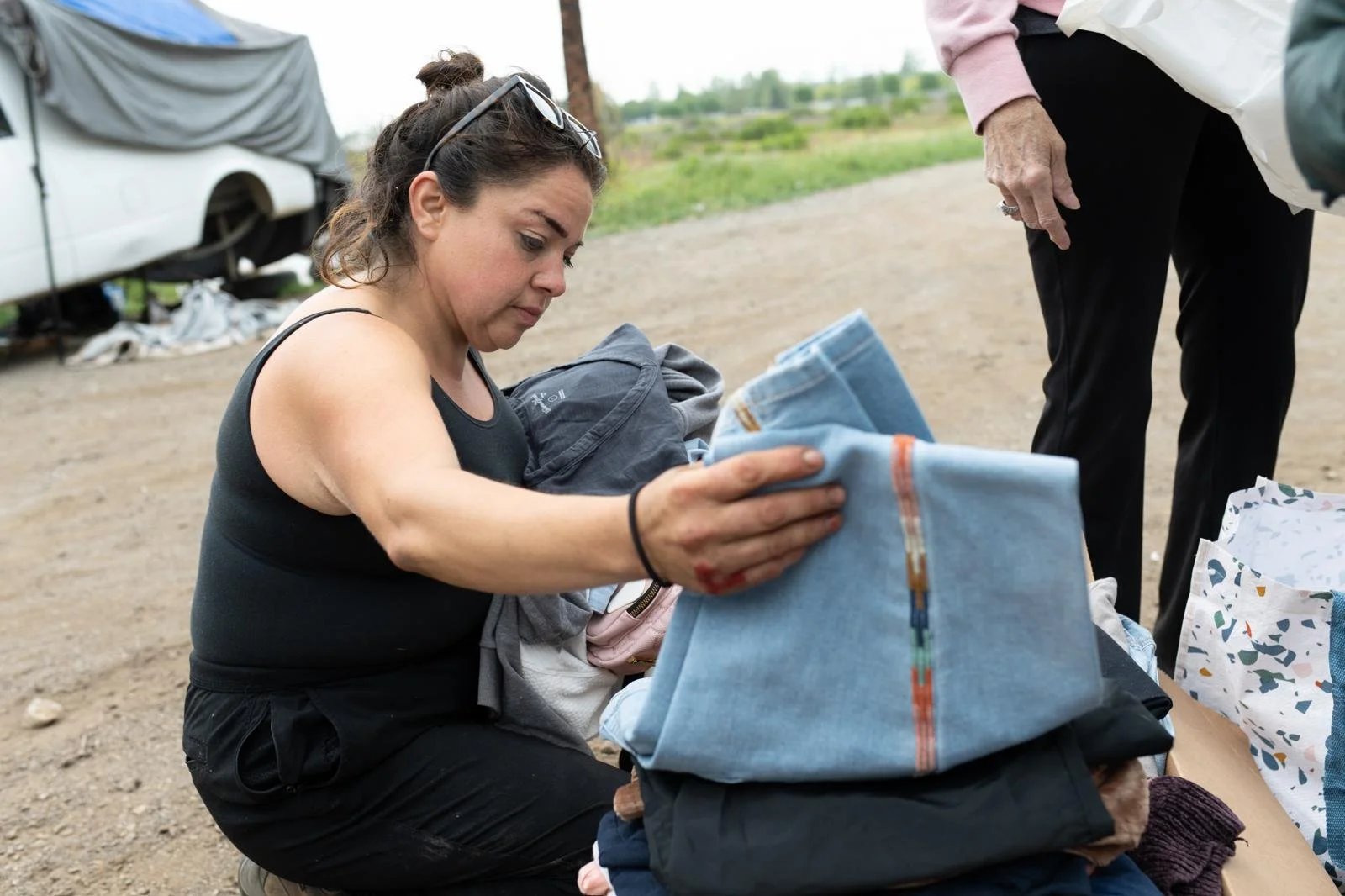 Woman folding a piece of clothing outdoors, with another person standing nearby and a vehicle in the background.