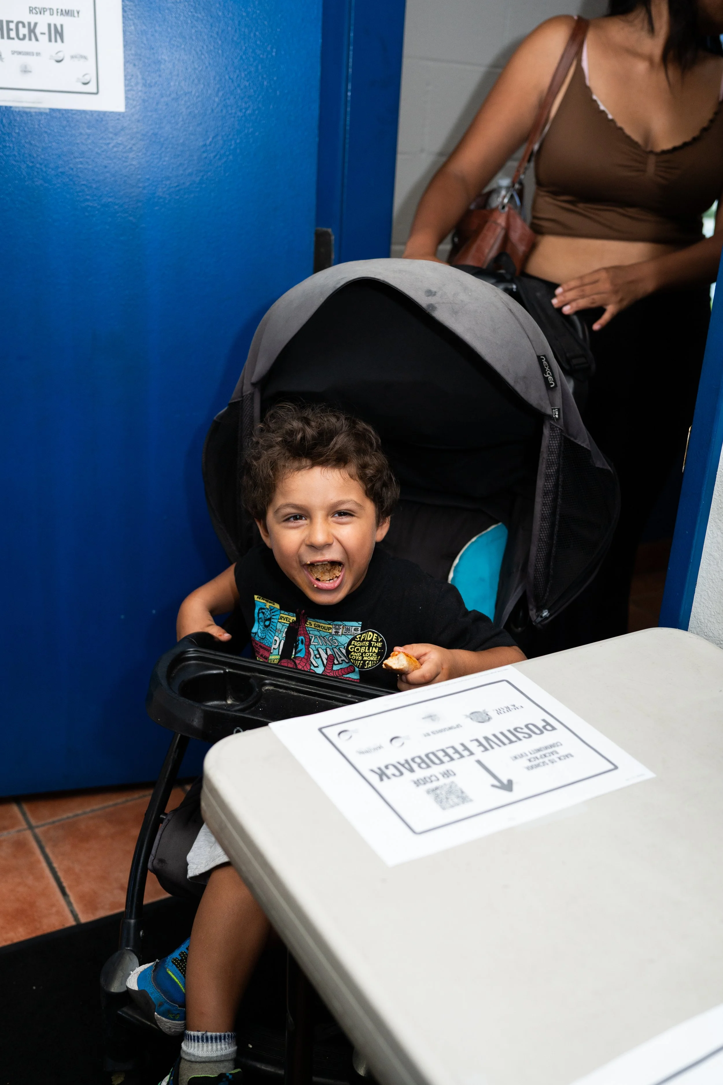 A smiling young boy sitting in a high chair at a petting zoo, holding a small animal, with a woman standing nearby, in an indoor setting with a blue wall and a registration table.