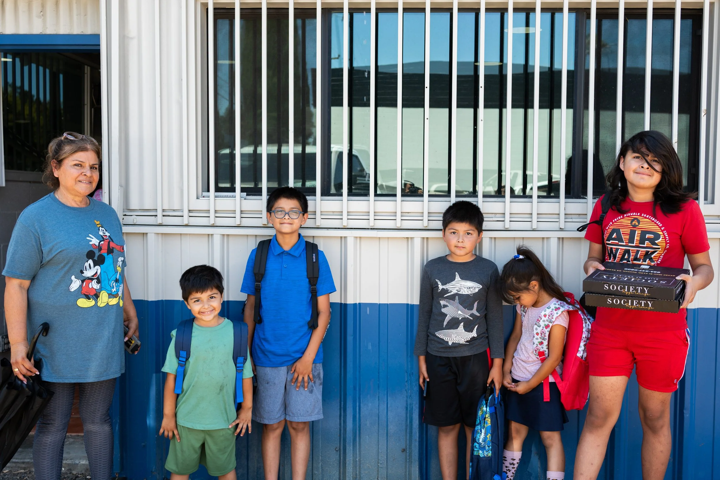 Group of children and two adults standing outside in front of a building with barred windows, smiling and carrying backpacks and boxes.