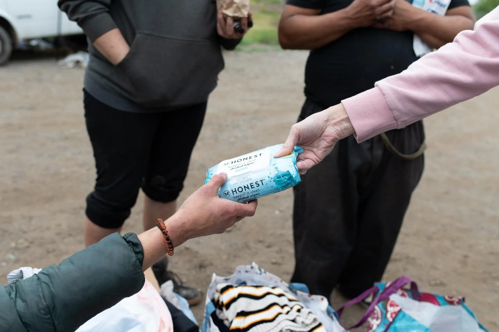 People exchanging packs of Honest alcohol wipes outdoors, with bags and clothing on the ground nearby.