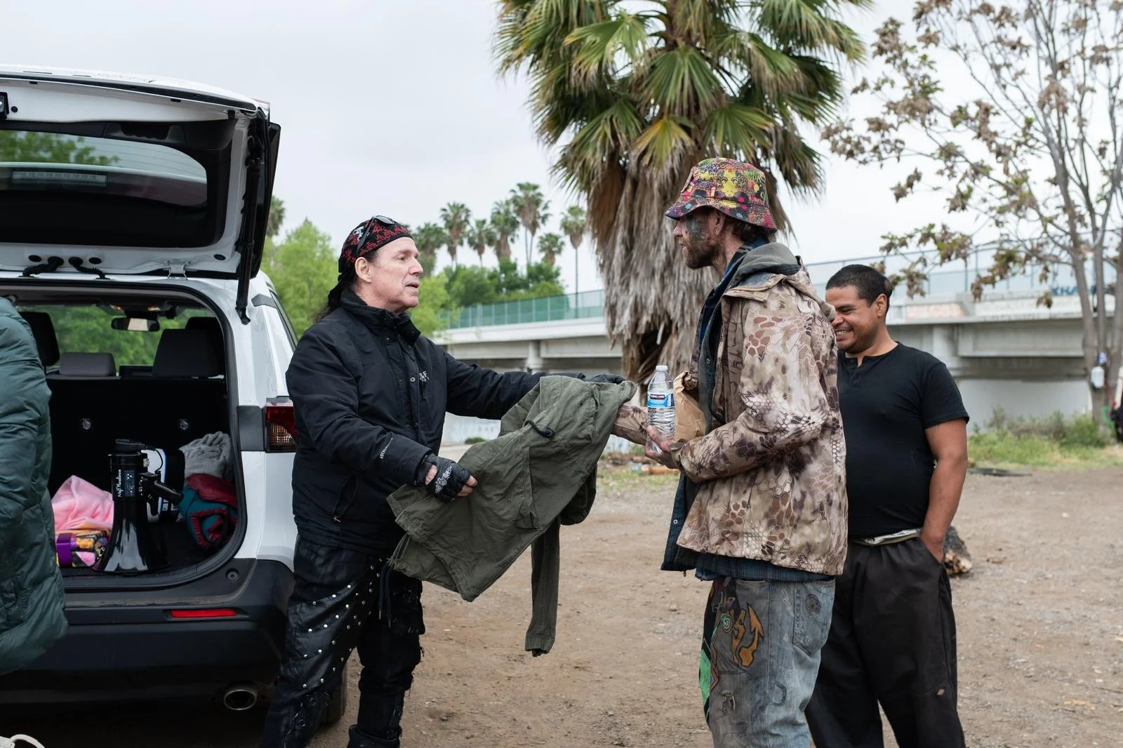 Person handing a water bottle and jacket to a man dressed in camouflage clothing, with two other men in black shirts in the background, in an outdoor area with trees and a highway overpass.