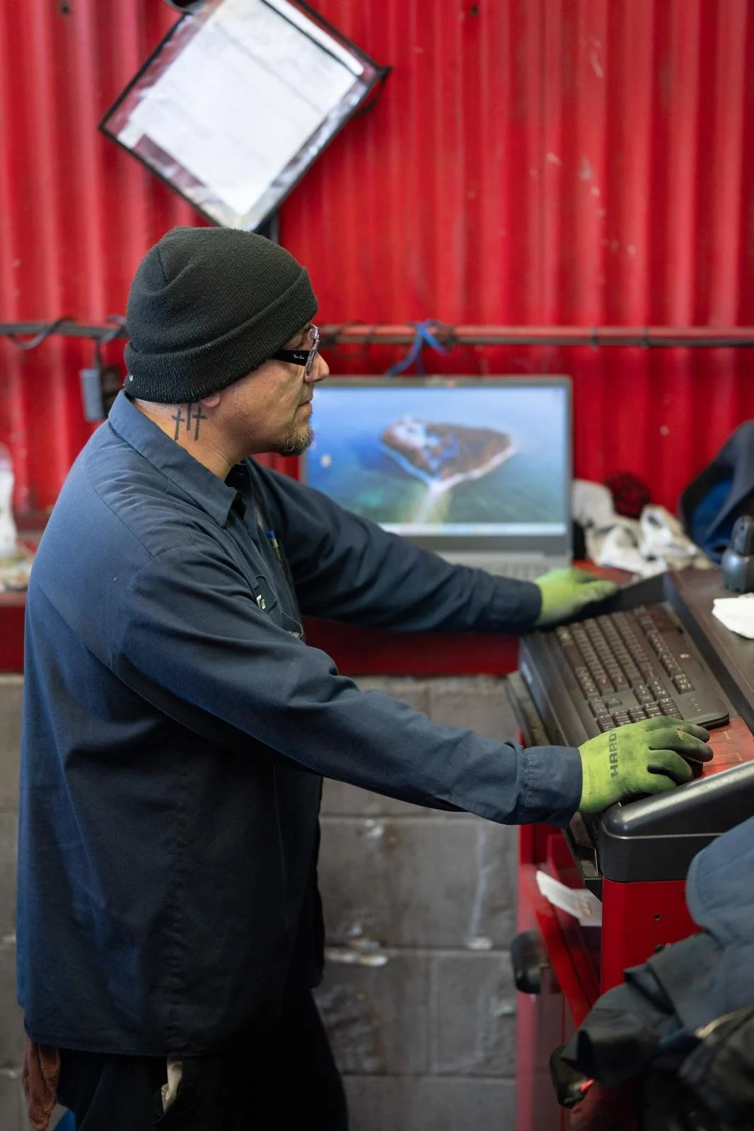 A man wearing a black beanie, glasses, a blue work shirt, and green gloves working at a computer workstation with a red background.