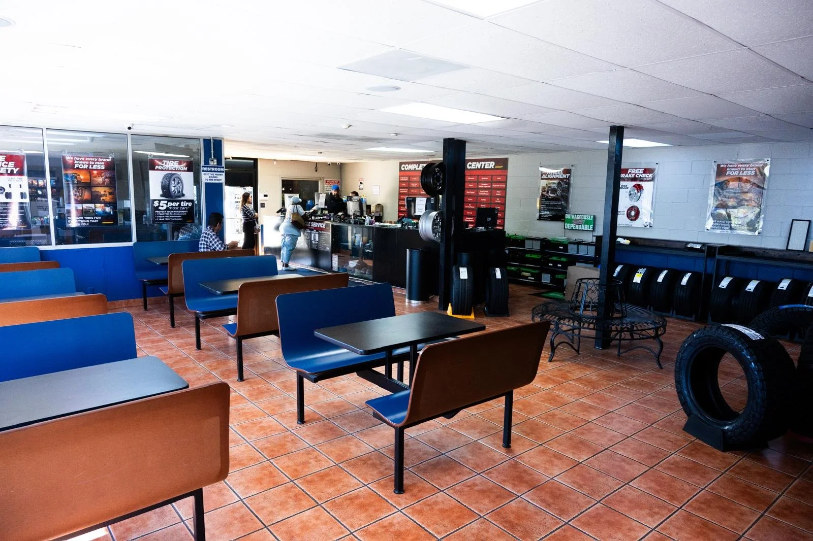 Interior of a tire shop or automotive service center with chairs and tables for waiting customers, shelves with tires, and a service counter with staff assisting customers.
