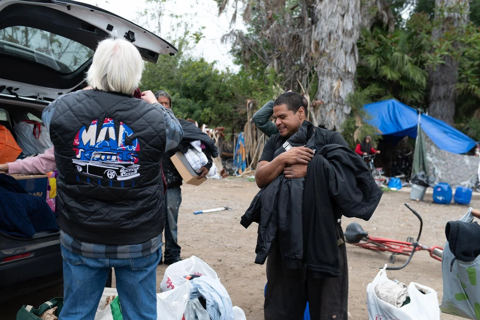 People gathering outdoors, unloading supplies from a vehicle, with trees and tents in the background.