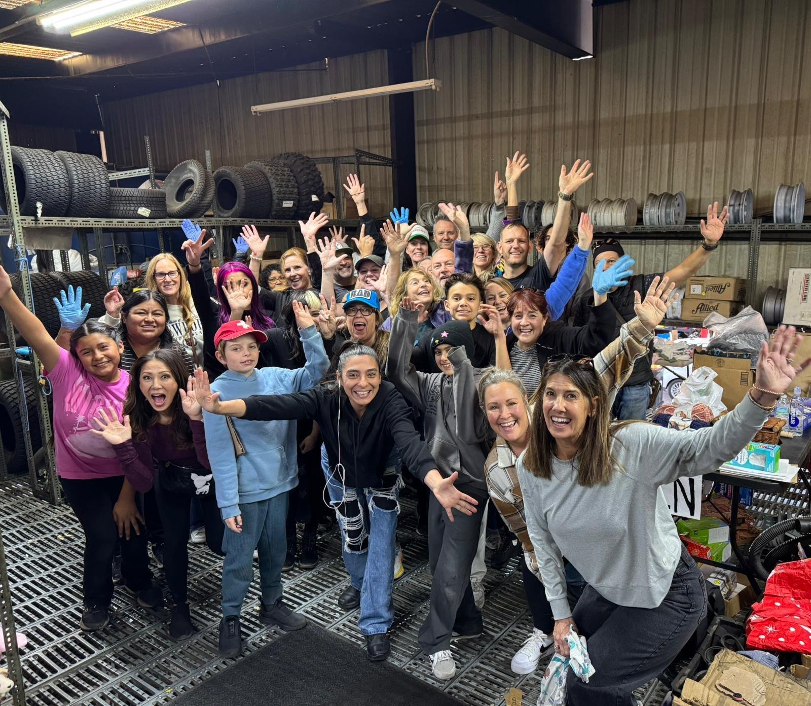 Group of smiling people, children and adults, inside a storage room or warehouse, celebrating and waving at the camera, with shelves stocked with tires and car rims behind them.