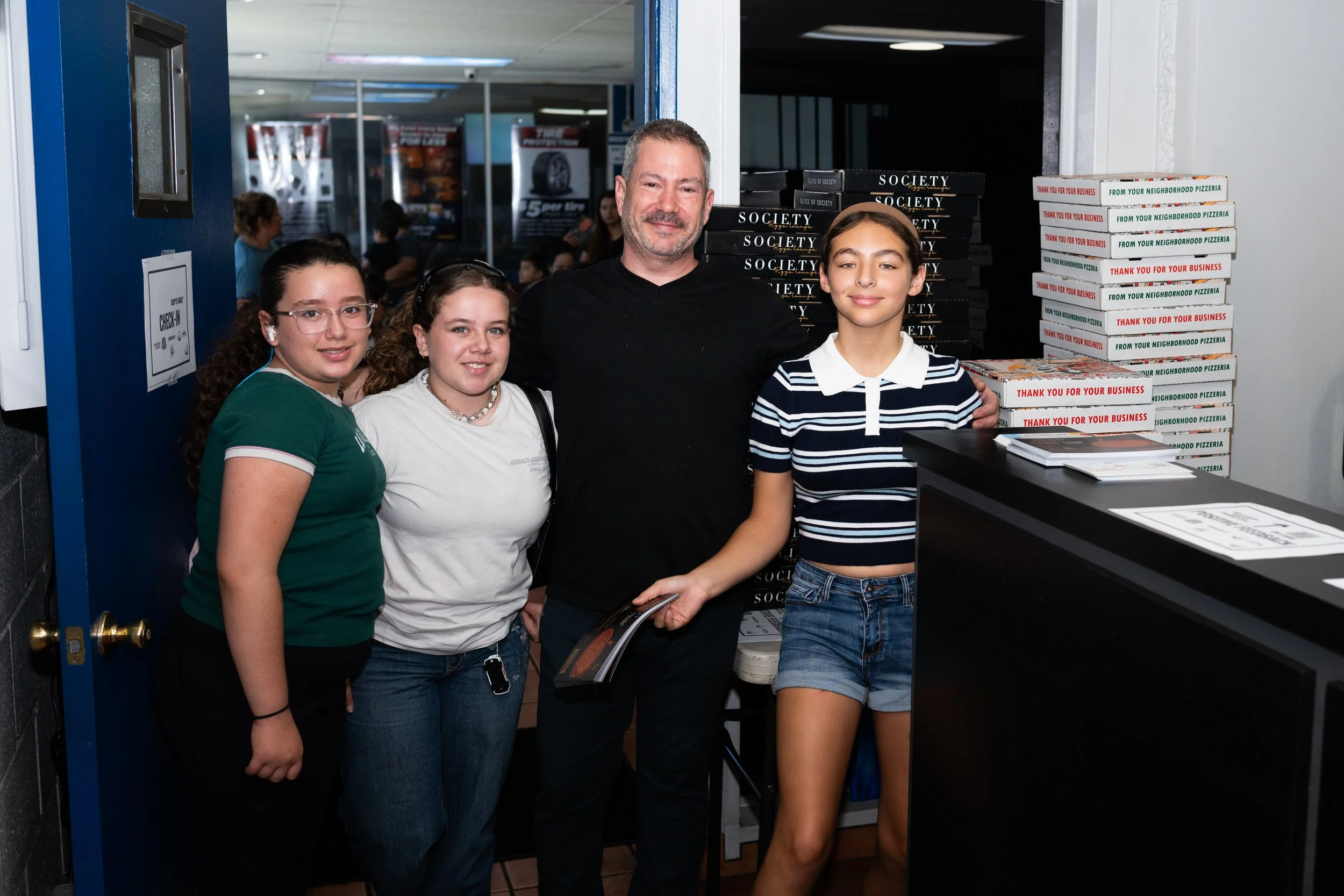 A group of four people, three young girls and an adult man, standing together inside a restaurant next to a stack of pizza boxes. The man is smiling, and the girls are looking at the camera. The background shows other customers and posters in the res