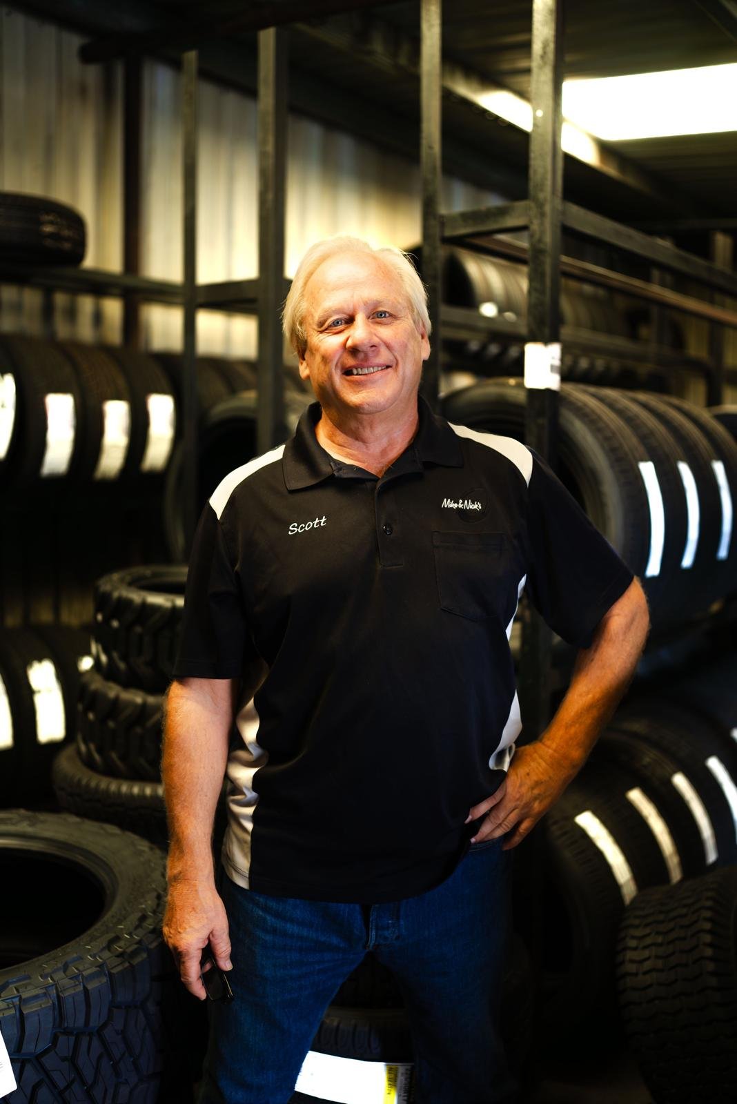 A smiling man wearing a black and white polo shirt with the name tag 'Scott' standing in an tire shop or warehouse with stacks of tires behind him.