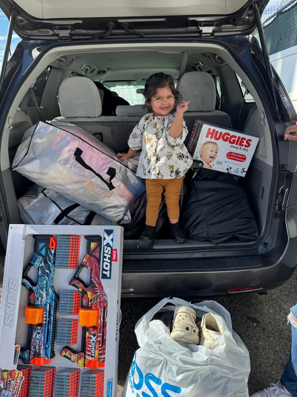 A young girl smiling and pointing inside the open trunk of a car, with various items including a box of Huggies diapers, a large silver bag, a boxed toy set, shoes, and other bags.