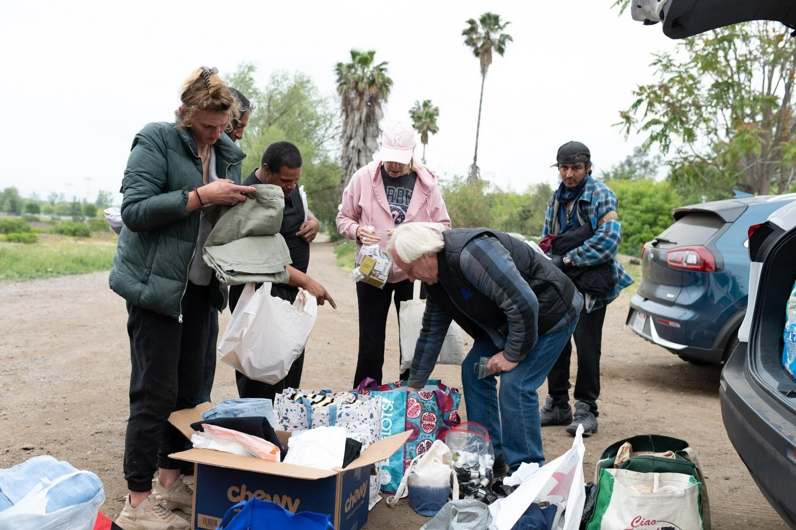 Group of six people sorting through supplies outdoors, with open car trunks and trees in the background.