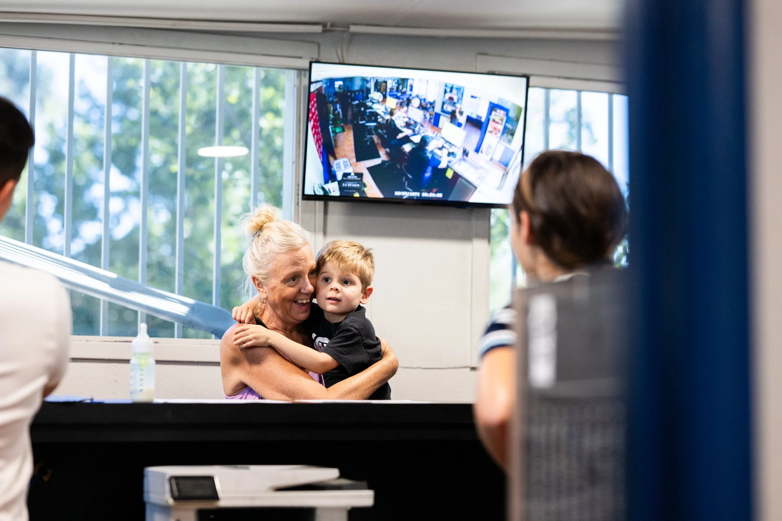 A woman holding a young boy hugging tightly in a room with a large window and a television screen showing multiple camera feeds in the background.