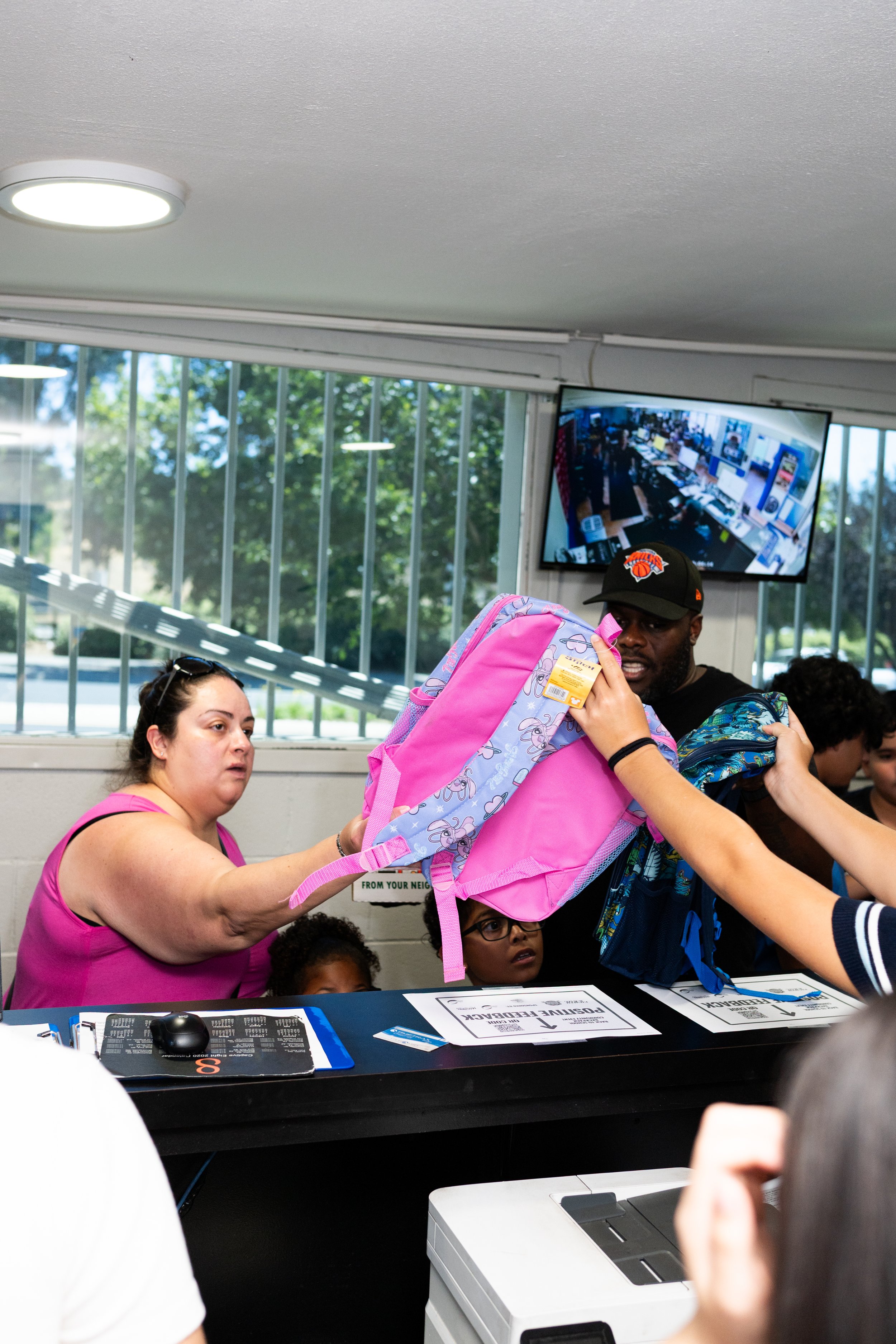 A group of people, including a woman in a pink top and a man wearing a New York Knicks cap, are at a polling station. The woman is handing a pink and purple backpack to the man. There are signs on the table for voting, and a monitor in the background