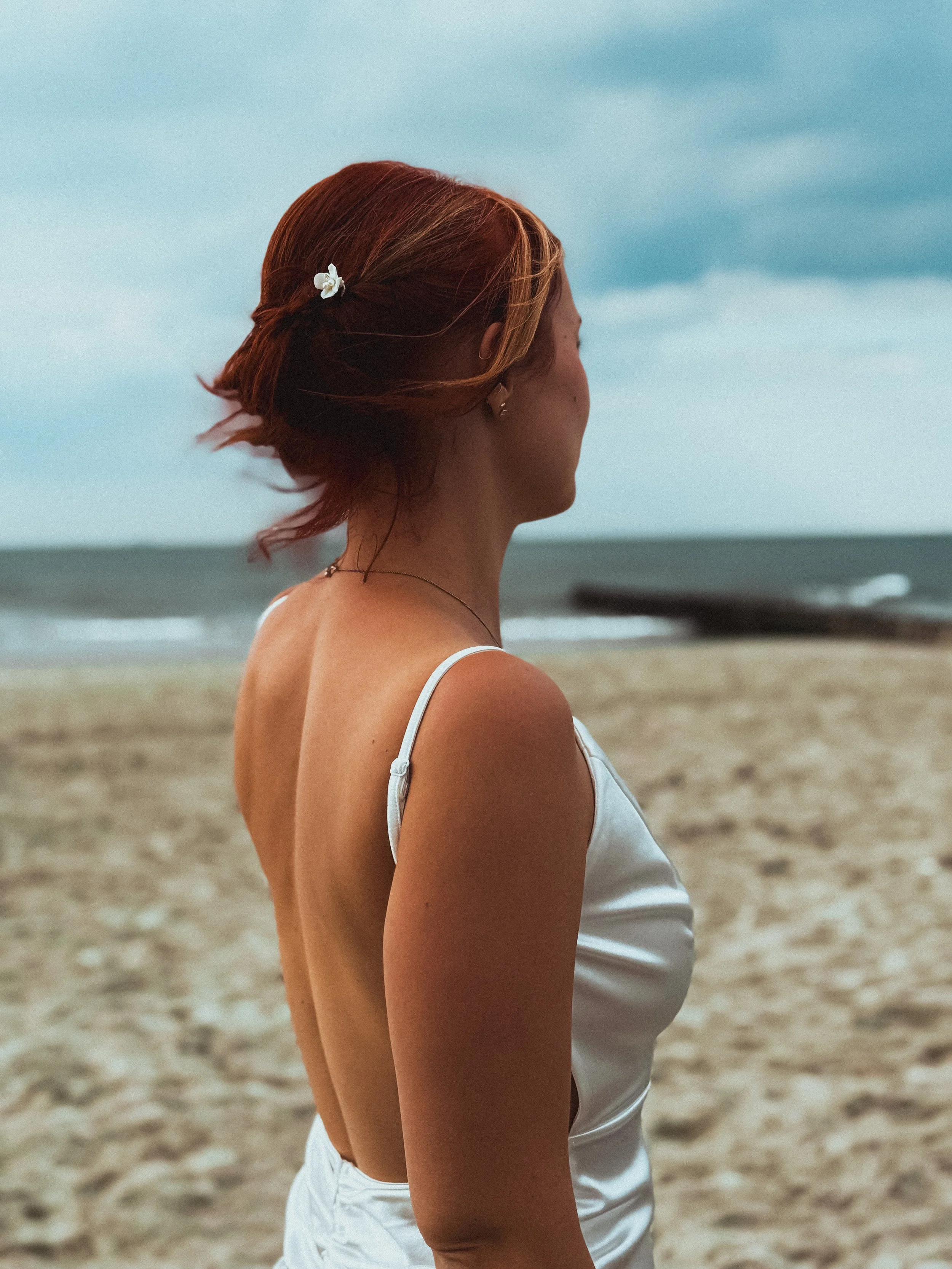 Braut mit roten Haaren und Haarschmuck steht am Strand und blickt aufs Meer