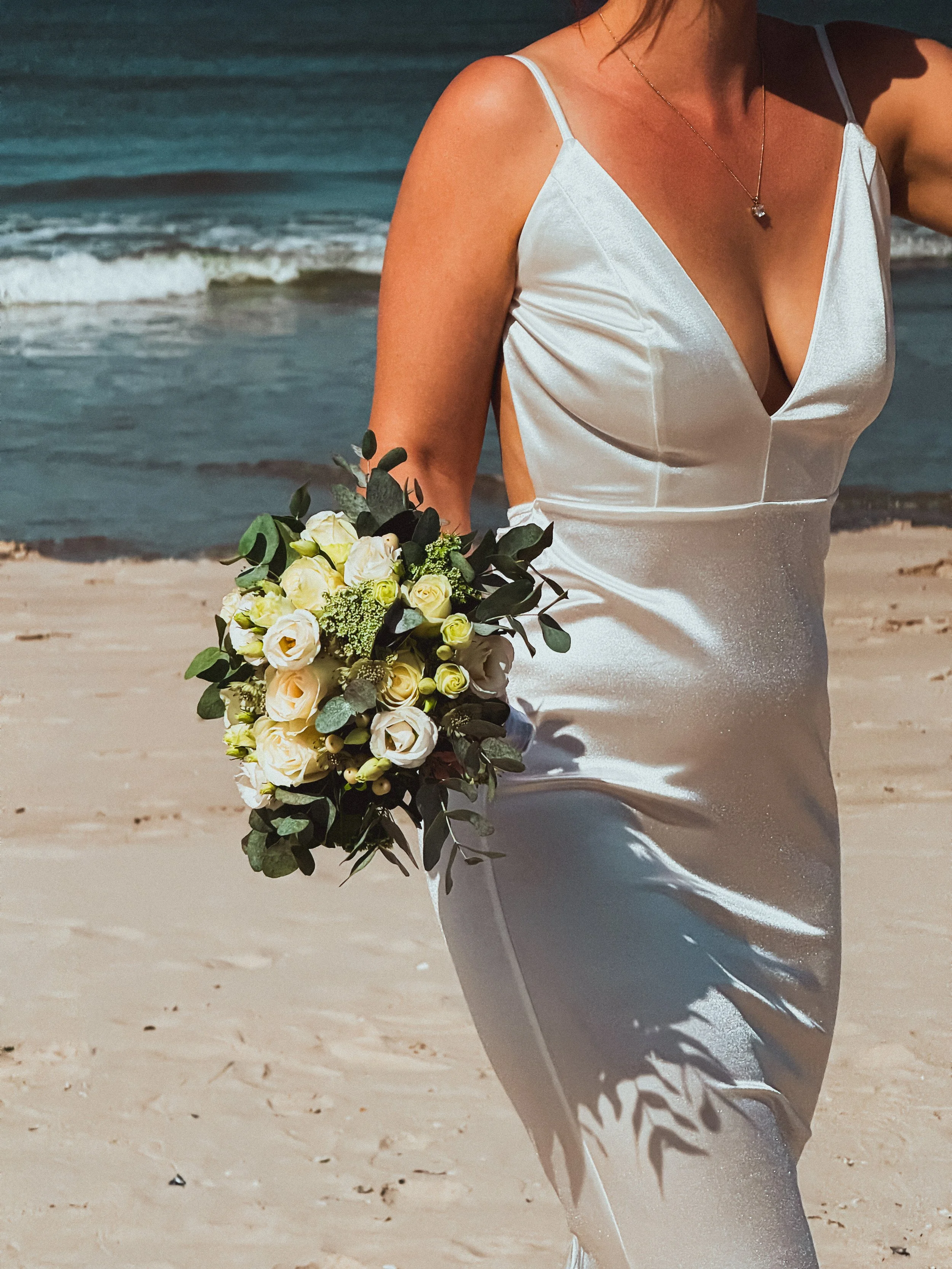 Braut im Satinhochzeitskleid mit weißem Blumenstrauß am Strand