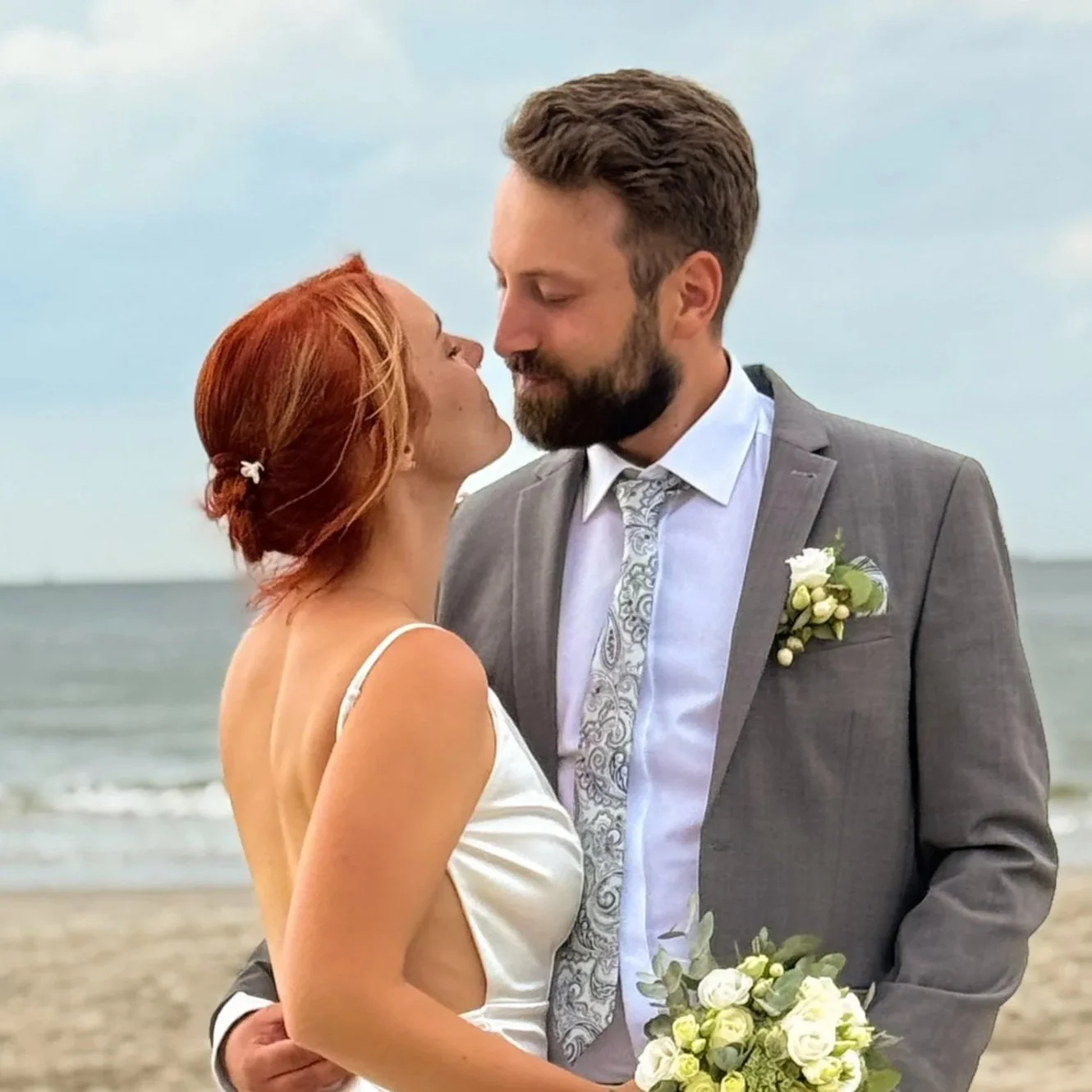 Brautpaar am Strand kurz vor einem Kuss, Braut mit roten Haaren, Bräutigam im grauen Anzug mit Blumenstrauß