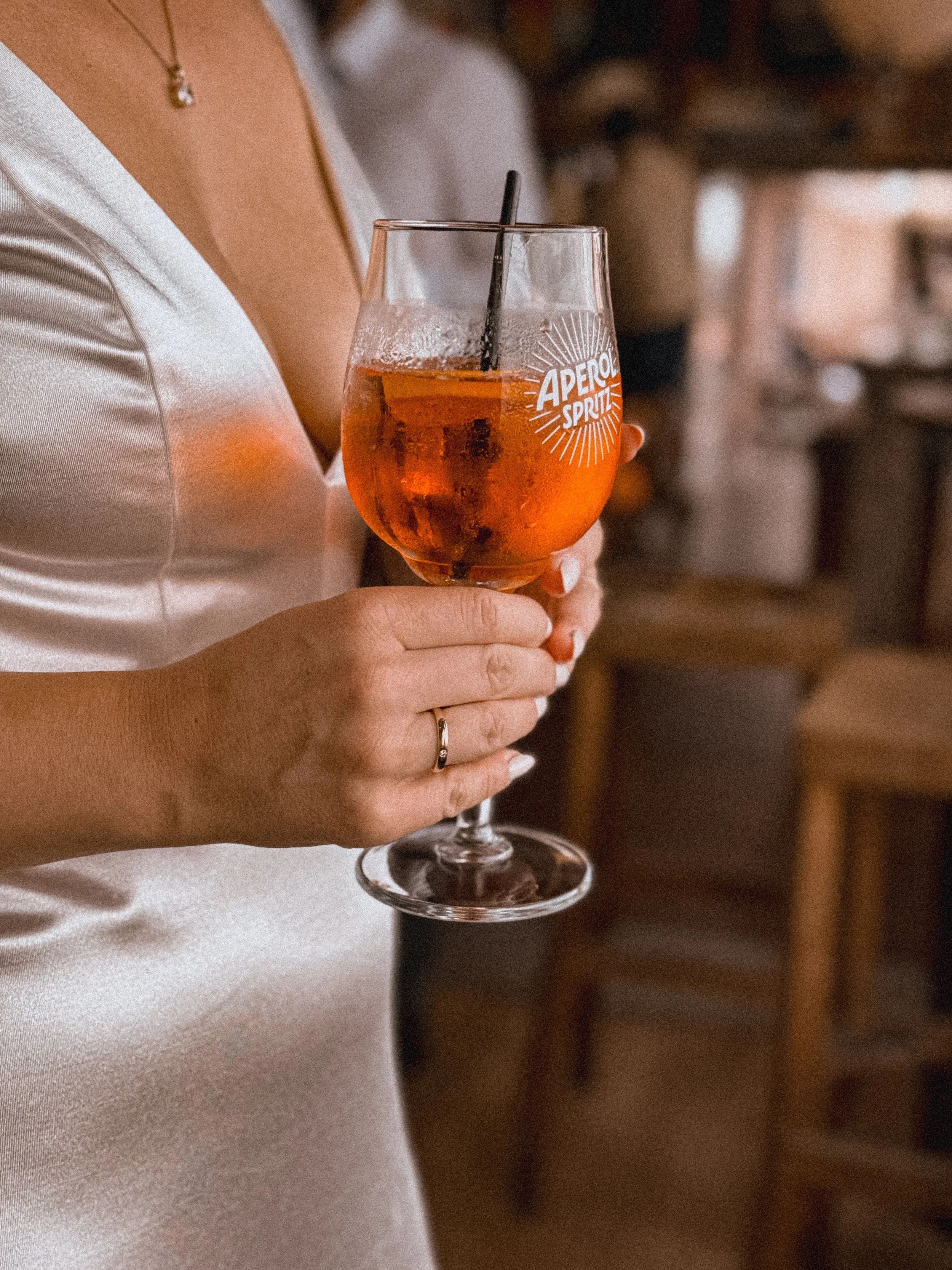 Braut hält ein Getränk im Glas, trägt Ring und Satinhochzeitskleid, in rustikaler Umgebung