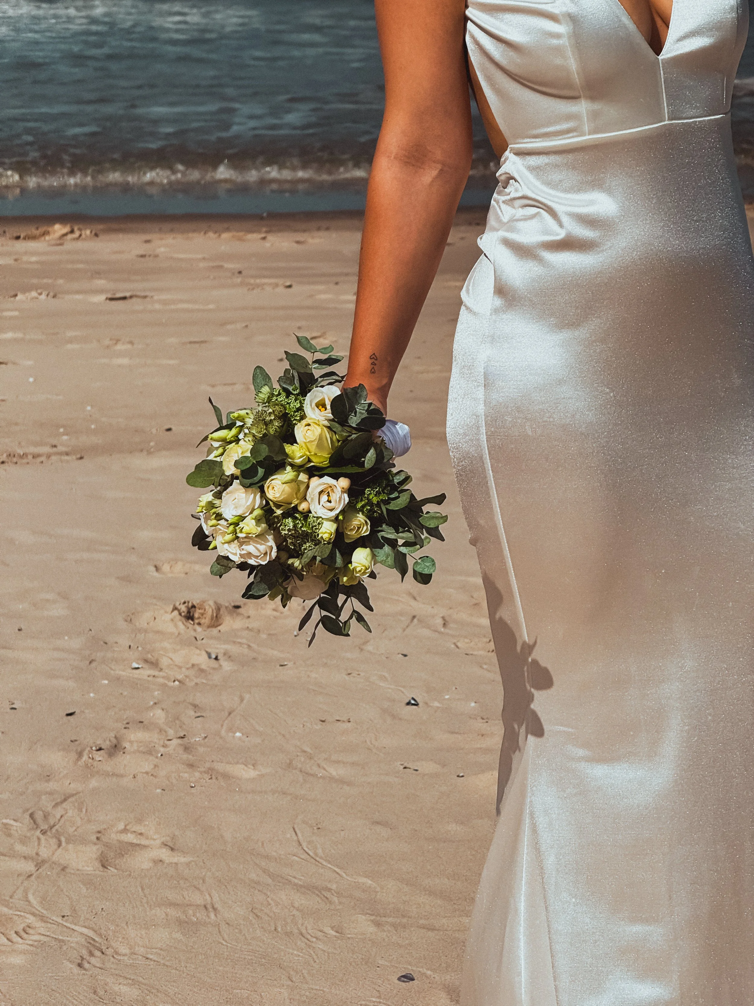 Braut im Satinhochzeitskleid mit Blumenstrauß aus weißen Rosen am Strand, Meer im Hintergrund
