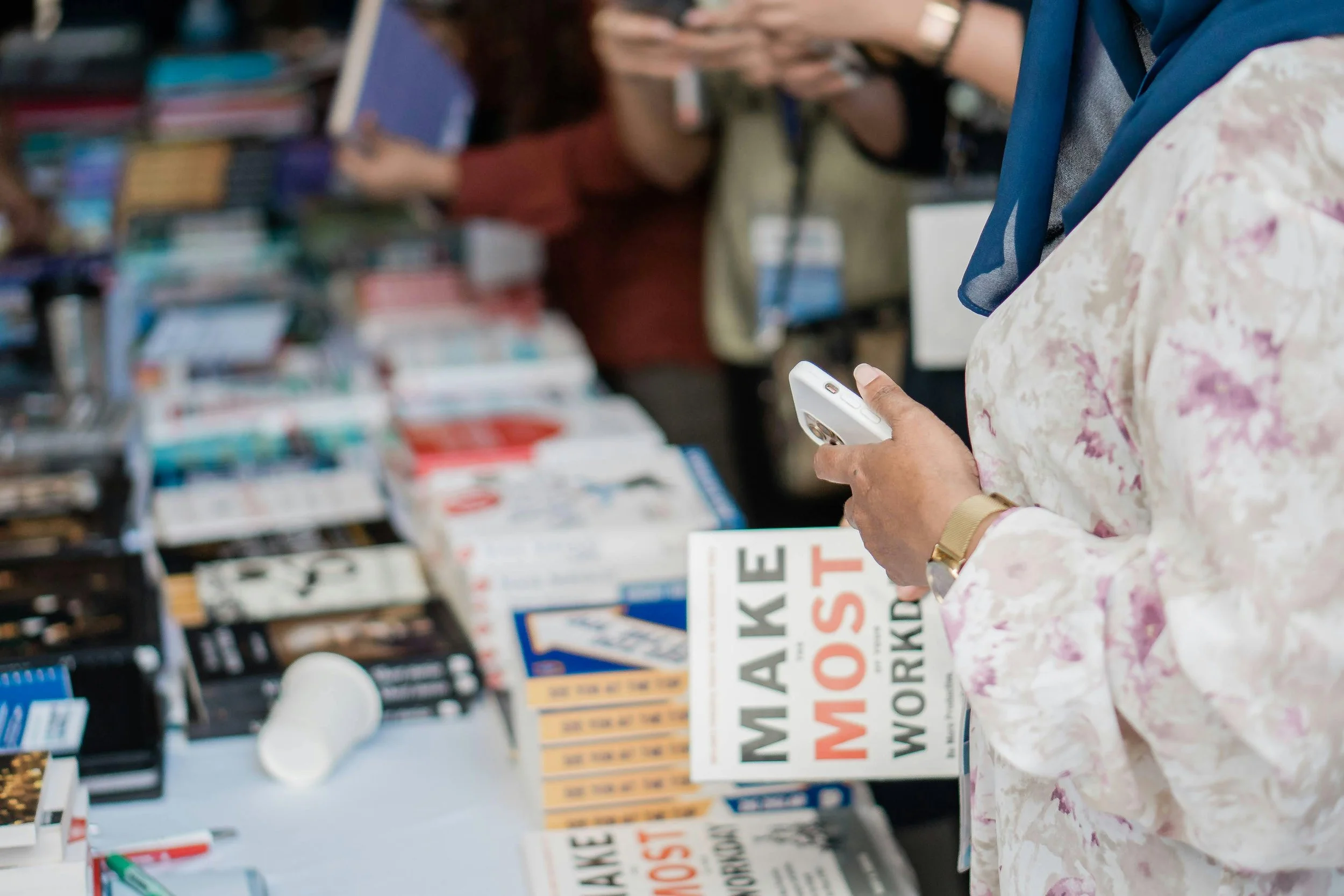 A close-up of a person wearing a floral shirt holding a smartphone near a table of books at a book fair or store. The table has multiple copies of the book "Make the Most."