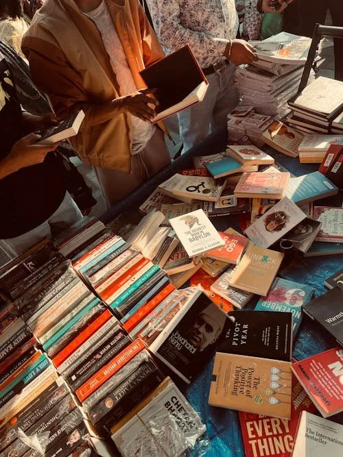 Books displayed on a table at a book market or store, with people browsing and holding books.