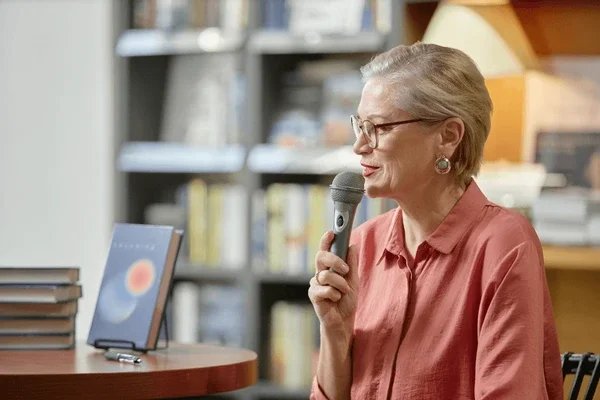 An elderly woman with short gray hair and glasses speaking into a microphone in a bookstore or library.