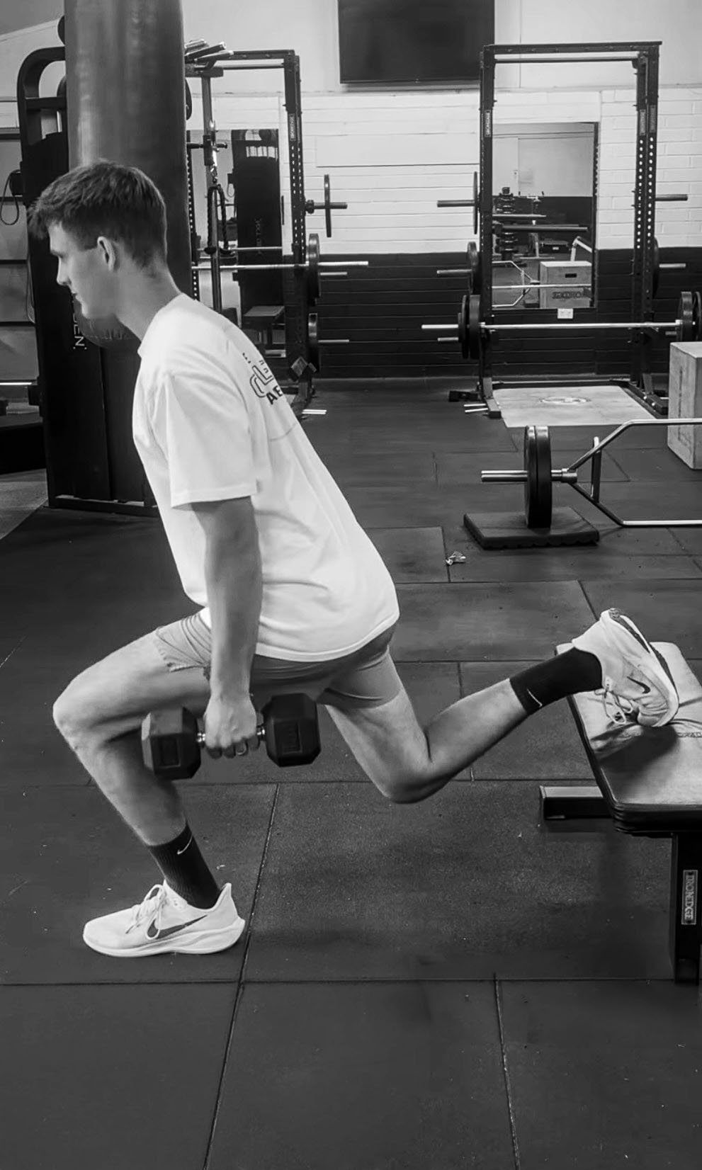 A man performing a Bulgarian split squat exercise in a gym, holding dumbbells in each hand, with one foot elevated on a bench behind him.