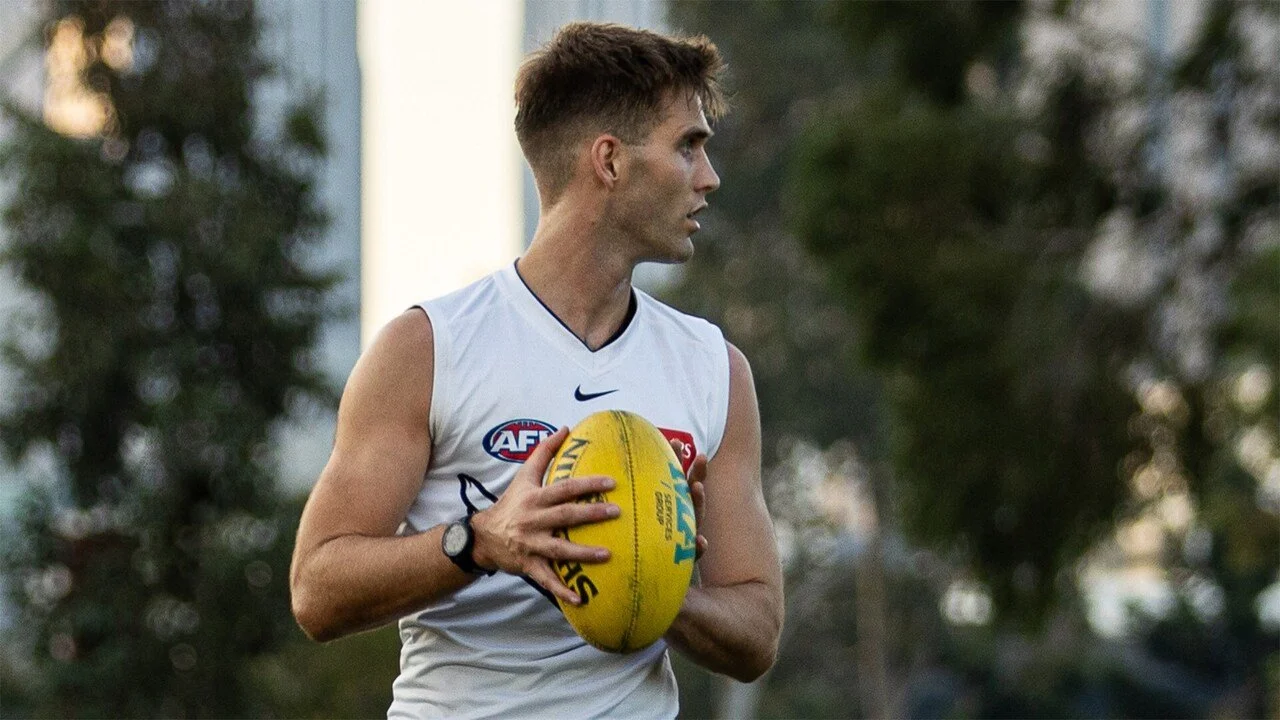 A young man in athletic gear holding an Australian rules football on a field, with trees and a building in the background.