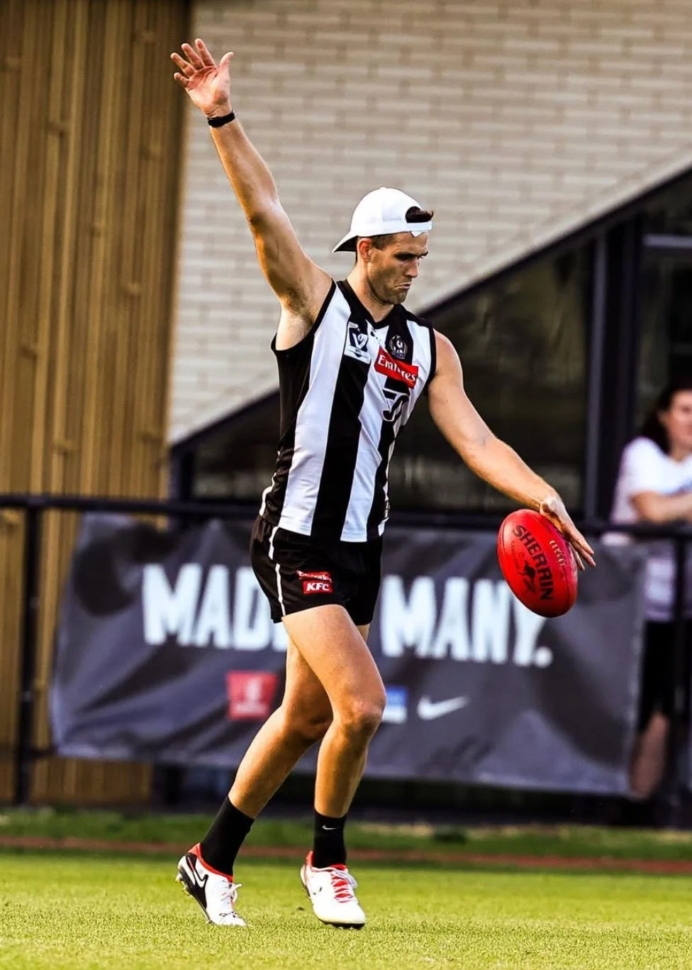 A man in Collingwood VFL club sports uniform, wearing a white cap backwards, is holding a red football and standing on a grassy field. His right arm is raised, and his expression is focused. Providing AFL coaching services in Victoria