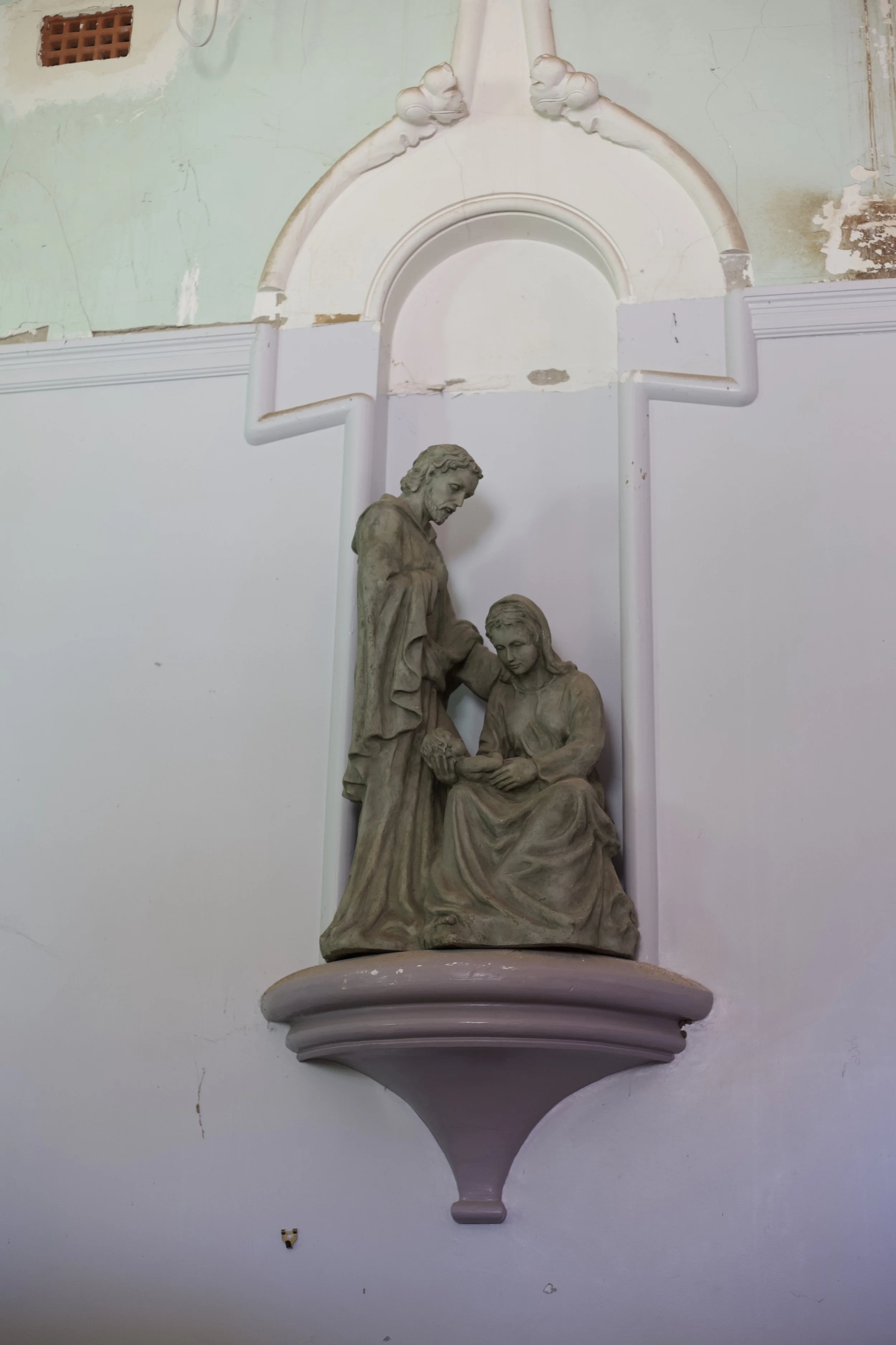 A religious or religious-style sculpture of three figures, with one standing and two seated, in a niche on a wall. The sculpture shows a man and two women, with the man placing his hands on the woman's shoulder as she holds a small object. The sculpture is placed above a decorative wall/niche and is made of a stone-like material.